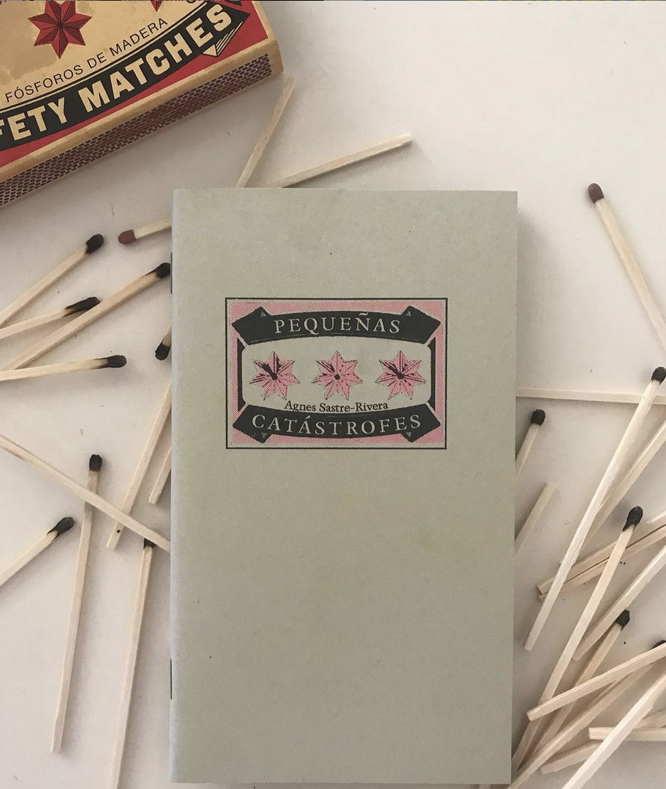 A collection of wooden matches with black and red heads scattered around a box of matches labeled in Spanish, reading 'pequeñas catástrofes' which translates to 'small catastrophes', with a book titled 'Pequeñas Catástrofes' by Agnes Sastres Rivera on a white surface.
