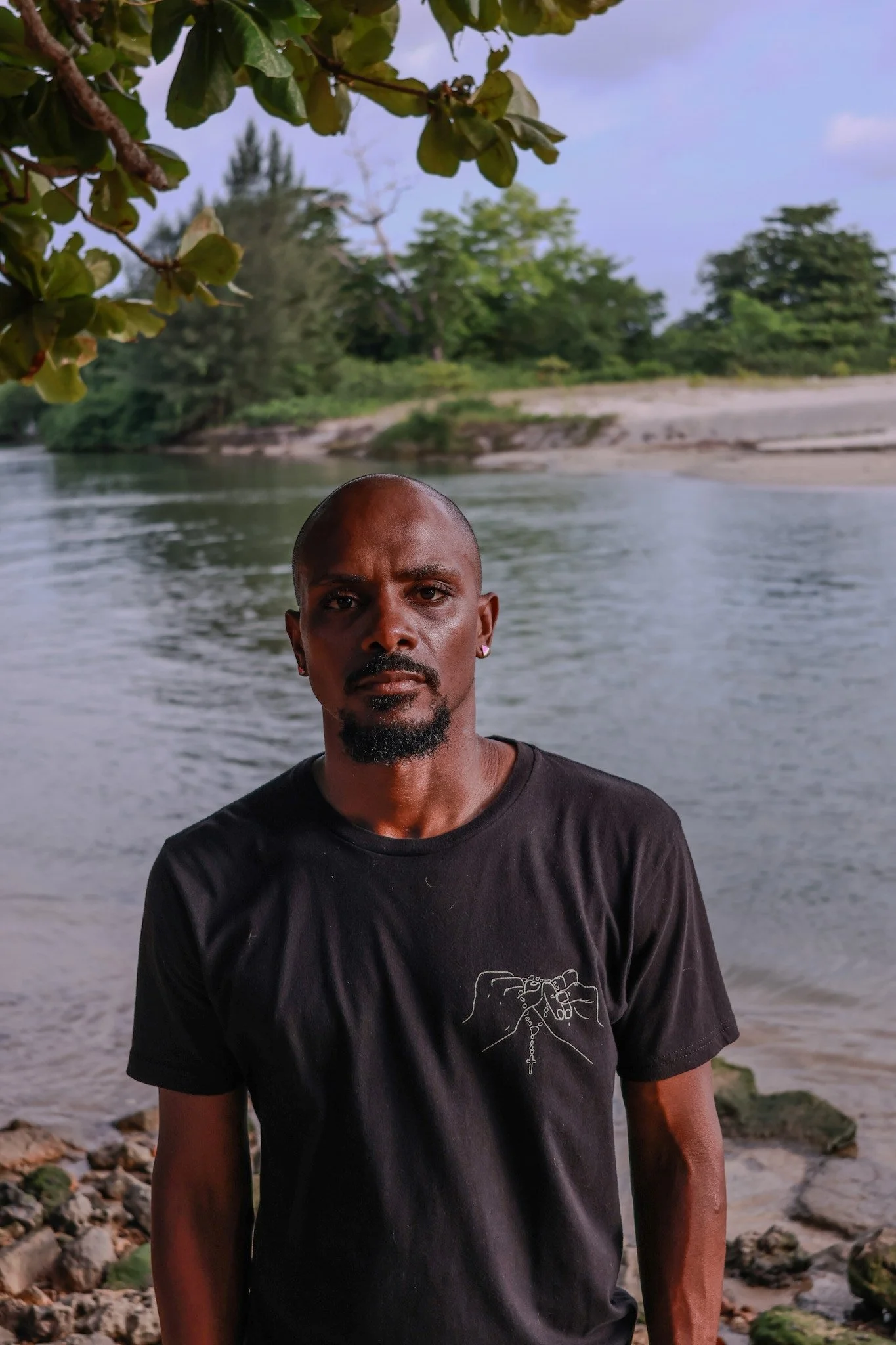 A man with a bald head and goatee standing by a river, looking directly at the camera. He is wearing a black T-shirt with a line drawing of hands holding a rosary. Trees and a sandy riverbank are in the background.