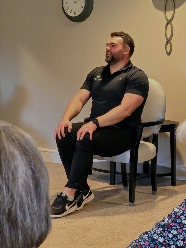 A man with a beard and short curly hair, wearing a black polo shirt and black pants, is sitting on a white cushioned chair in a room with beige walls. He is smiling and appears to be speaking or listening to someone. In the background, there is a wall clock and a decorative wall piece with circles. Several people with gray and white hair are visible in the foreground, suggesting an audience.