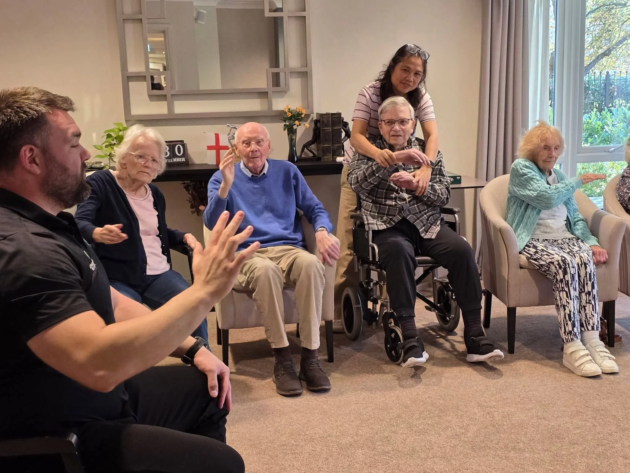 Group of elderly people participating in a seated activity at a care home or community center, with one caregiver assisting and engaging them in conversation or a game.