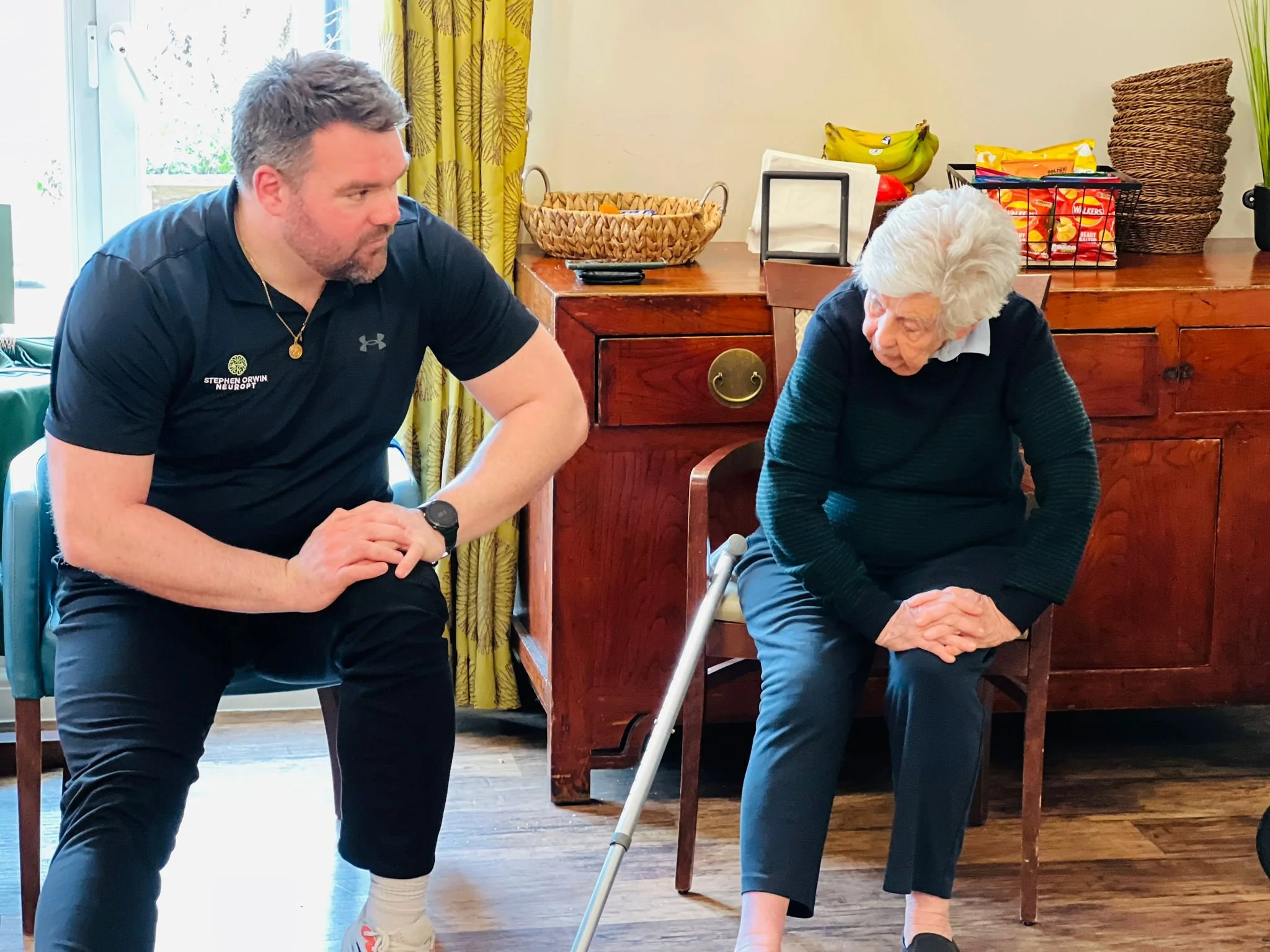 A man kneeling and a woman seated in a chair with a cane in her hand, inside a room with wooden floors and a wooden cabinet, both engaging in conversation.