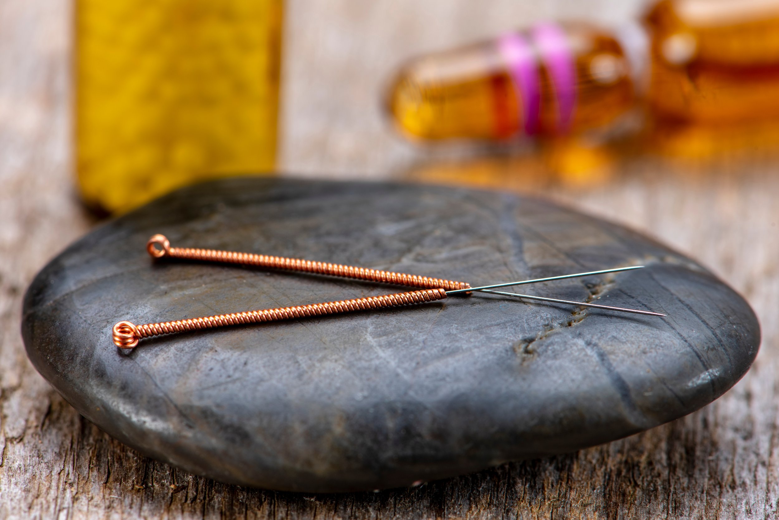 Acupuncture needles on a smooth stone with blurred background, including a yellow bottle and capsule.