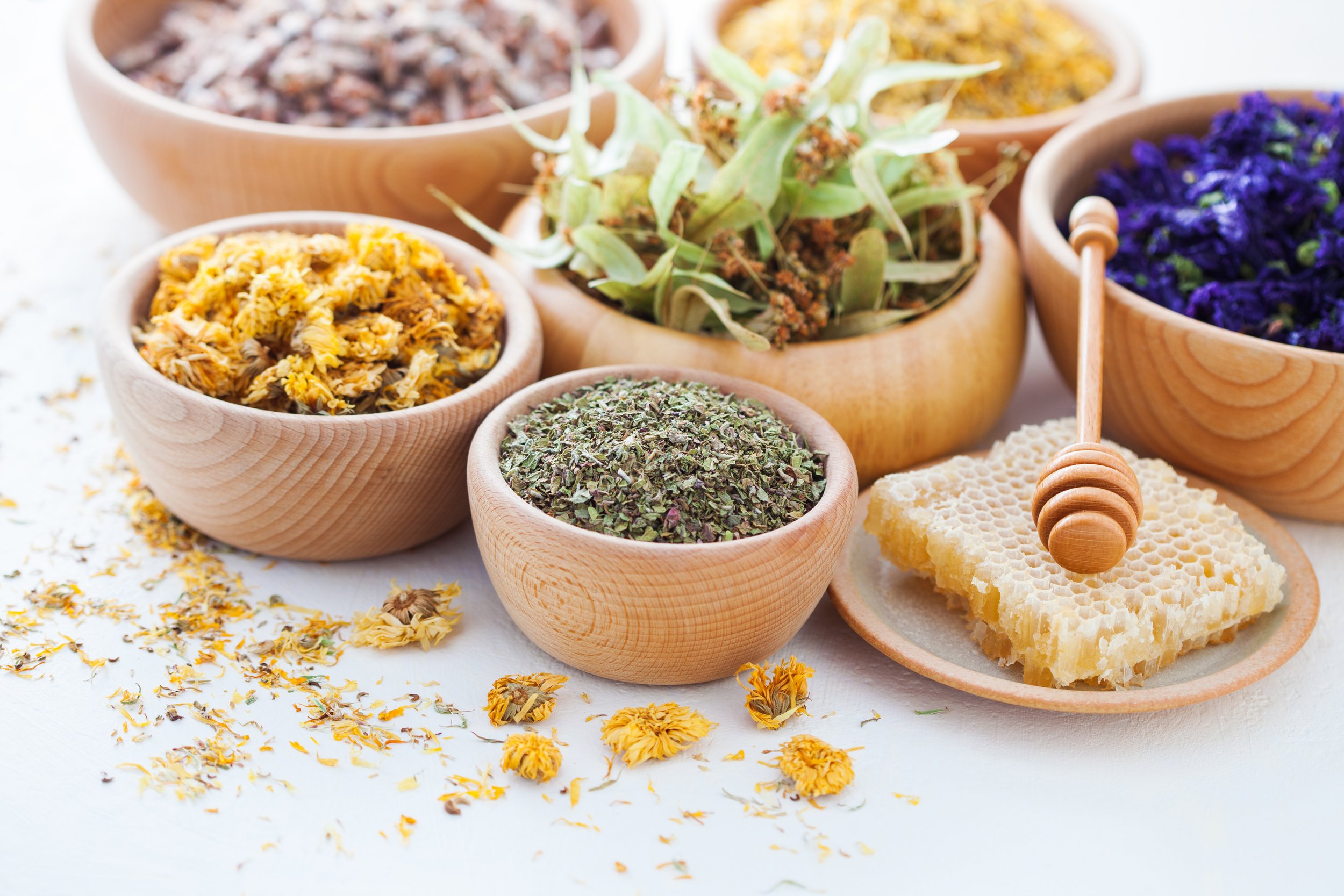Various dried herbs and flowers in wooden bowls with honeycomb on a plate and a honey dipper.