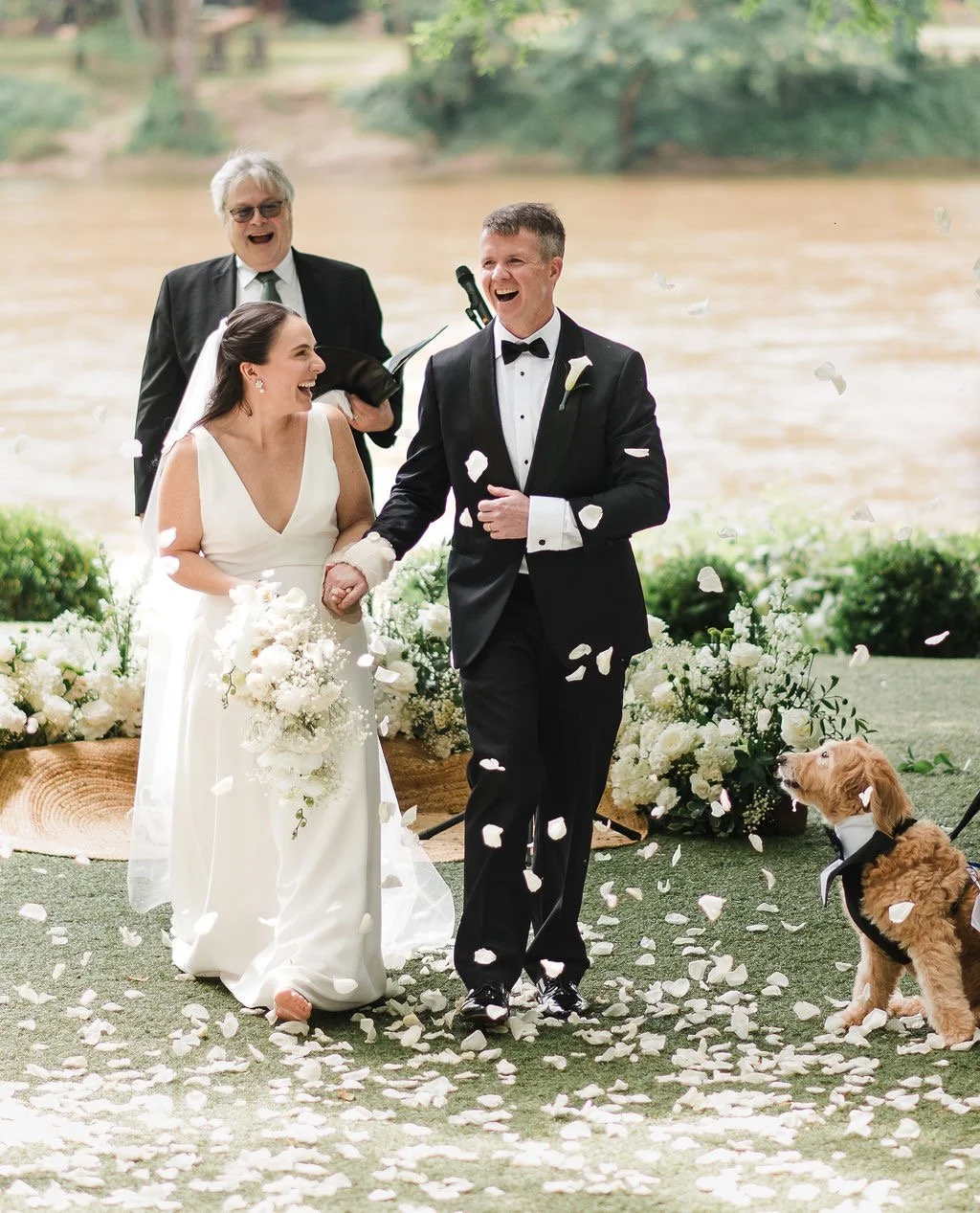 A bride and groom holding hands and smiling during their wedding ceremony outdoors, with an officiant, dog, and floral decorations, as flower petals fall around them by a river.