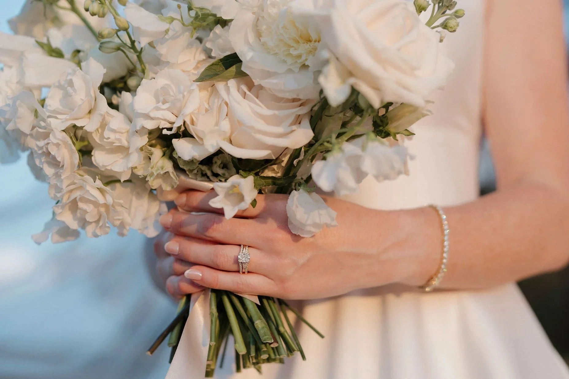 A bride holding a bouquet of white flowers, with her left hand visible, wearing a diamond engagement ring and a bracelet.
