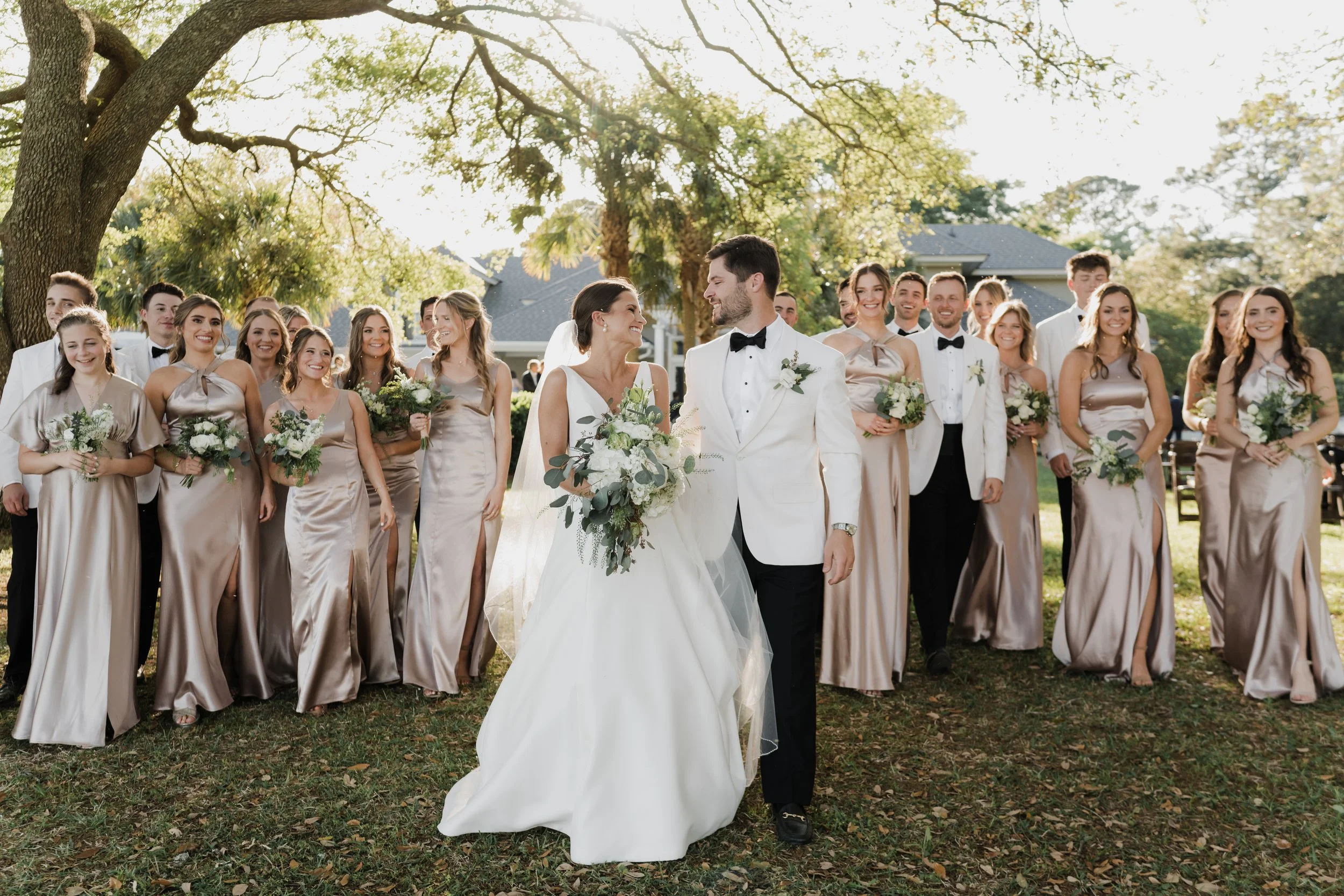 A bride and groom walking together and smiling at each other during their wedding ceremony outdoors, surrounded by bridesmaids and groomsmen holding bouquets, under a large tree on a sunny day.