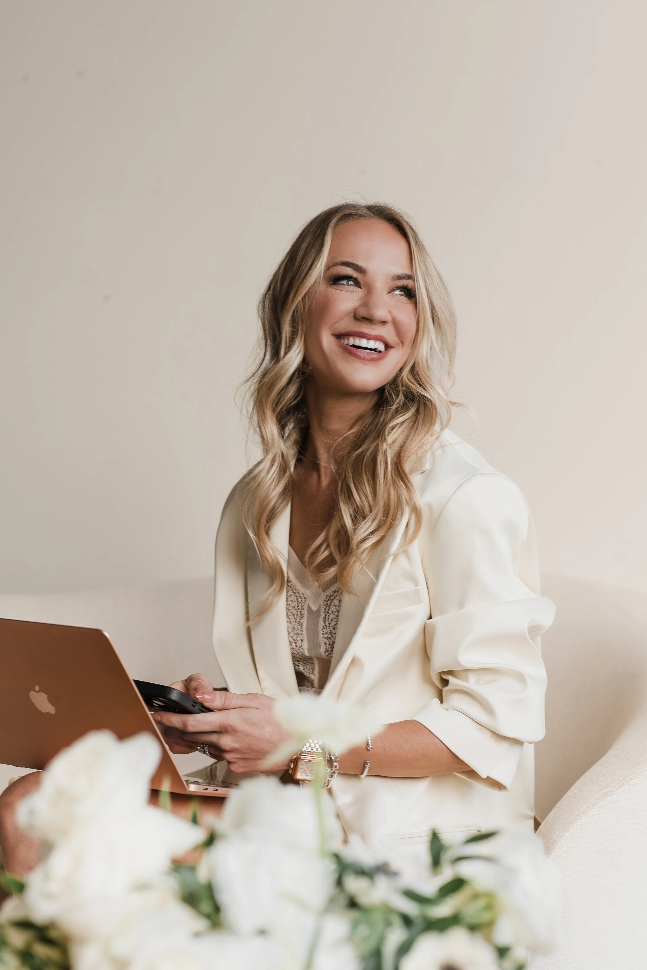 A smiling woman with wavy blonde hair, wearing a white blazer, sitting on a white couch, holding a smartphone, with a laptop in front of her and white flowers in the foreground.