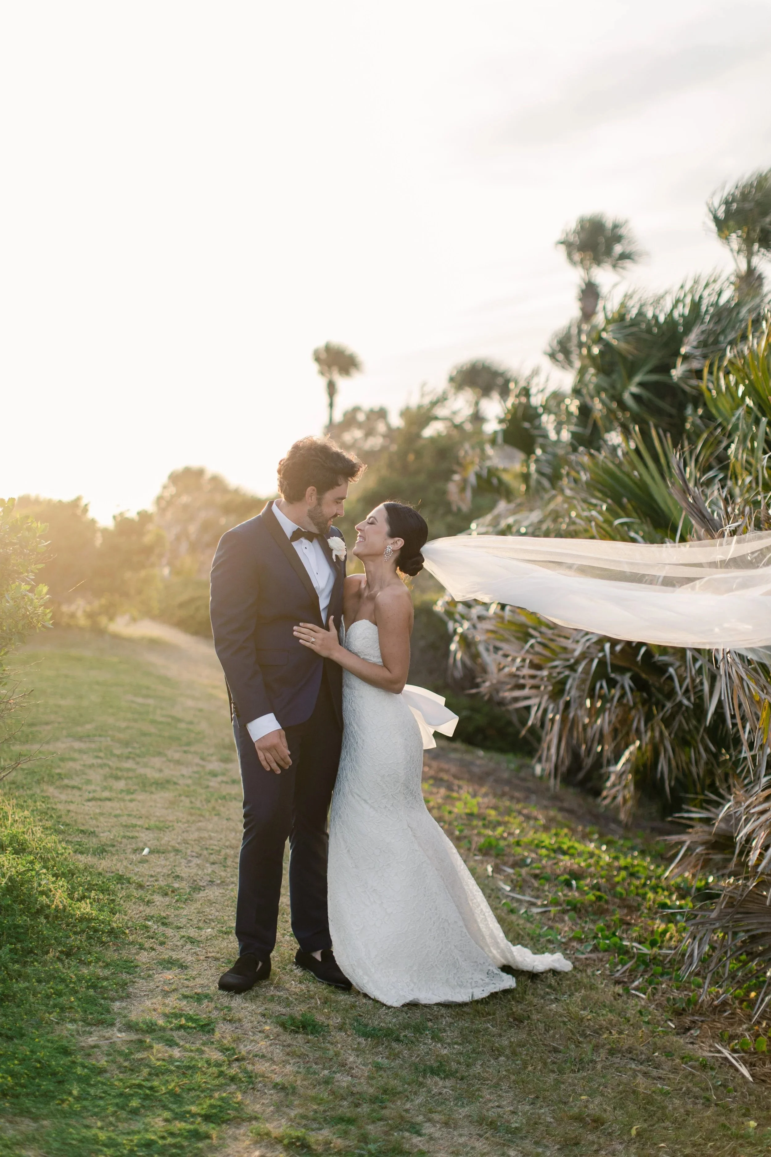A bride and groom standing close together outdoors during sunset, with lush greenery and palm trees in the background. The bride is wearing a white strapless wedding gown with a long train and veil, and the groom is in a black tuxedo. They are smiling and gazing into each other's eyes.
