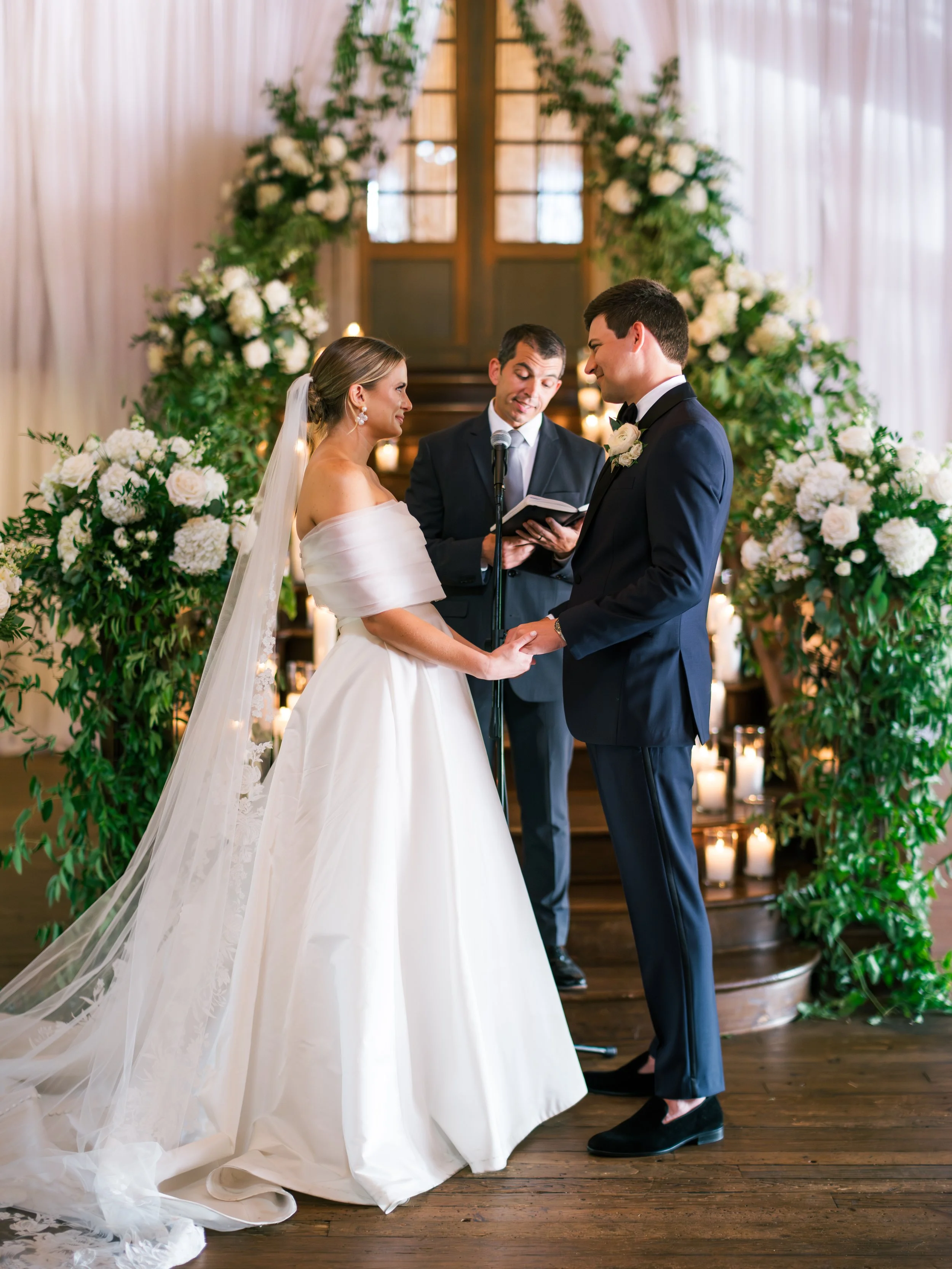 Bride and groom holding hands during their wedding ceremony, with officiant reading vows, surrounded by floral arrangements and candles.