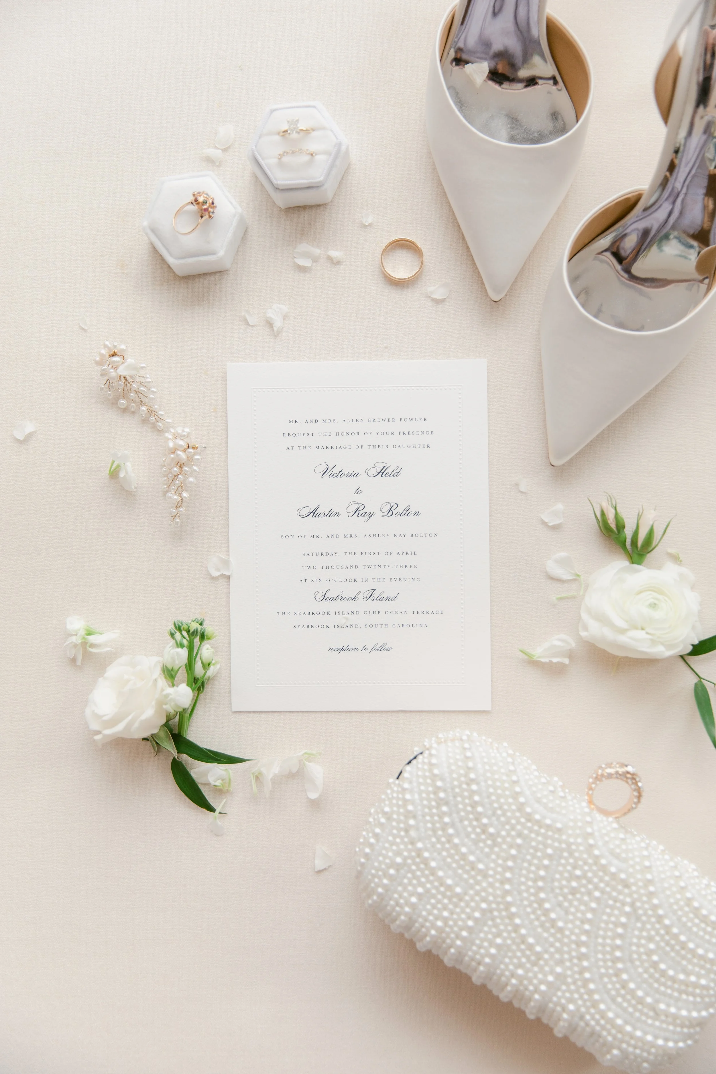 Wedding invitation surrounded by white flowers, pearl jewelry, white high heels, two glasses of iced coffee, a white beaded clutch purse, and a ring on an elegant table setting.