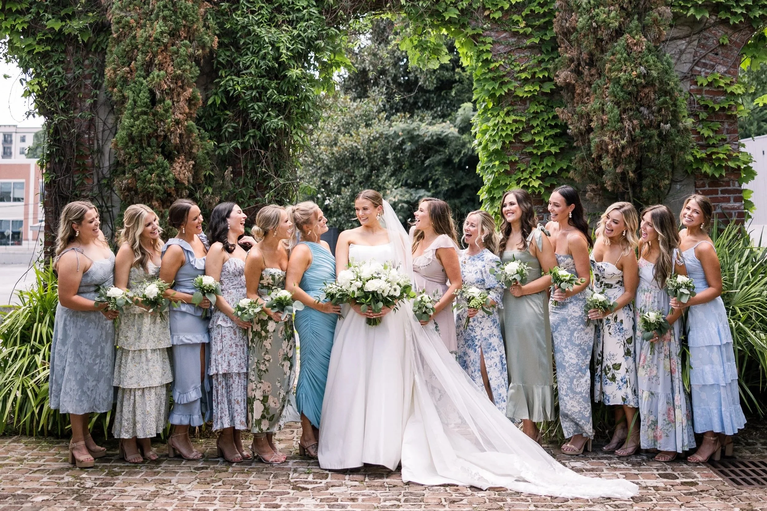 Bride surrounded by bridesmaids in soft pastel dresses, smiling amid greenery — timeless Southern bridal party style.