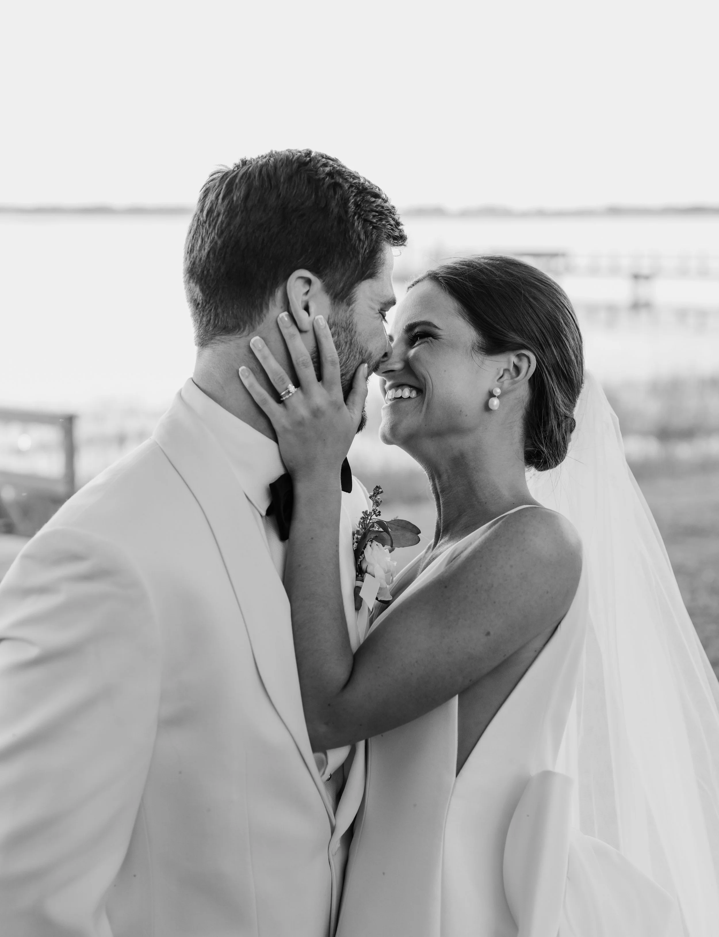 Black and white photo of a bride and groom smiling and touching noses, standing outdoors with water and sky in the background.
