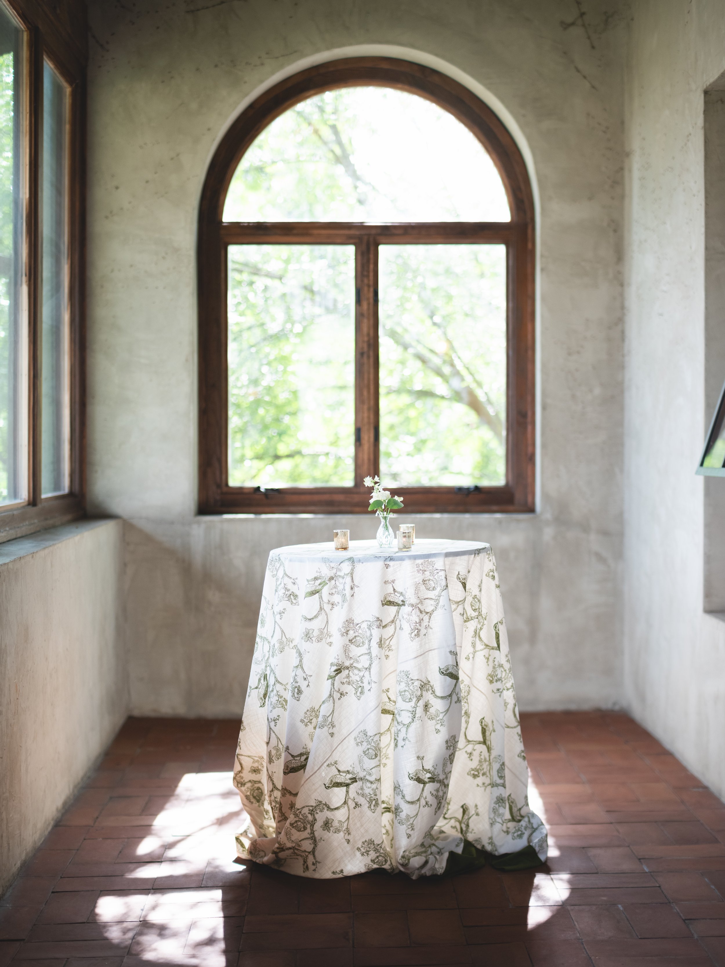 A round table covered with a cream-colored tablecloth with green floral patterns, a small vase with white flowers, and three small candles, set in front of a large arched window with wood trim, letting in natural light.