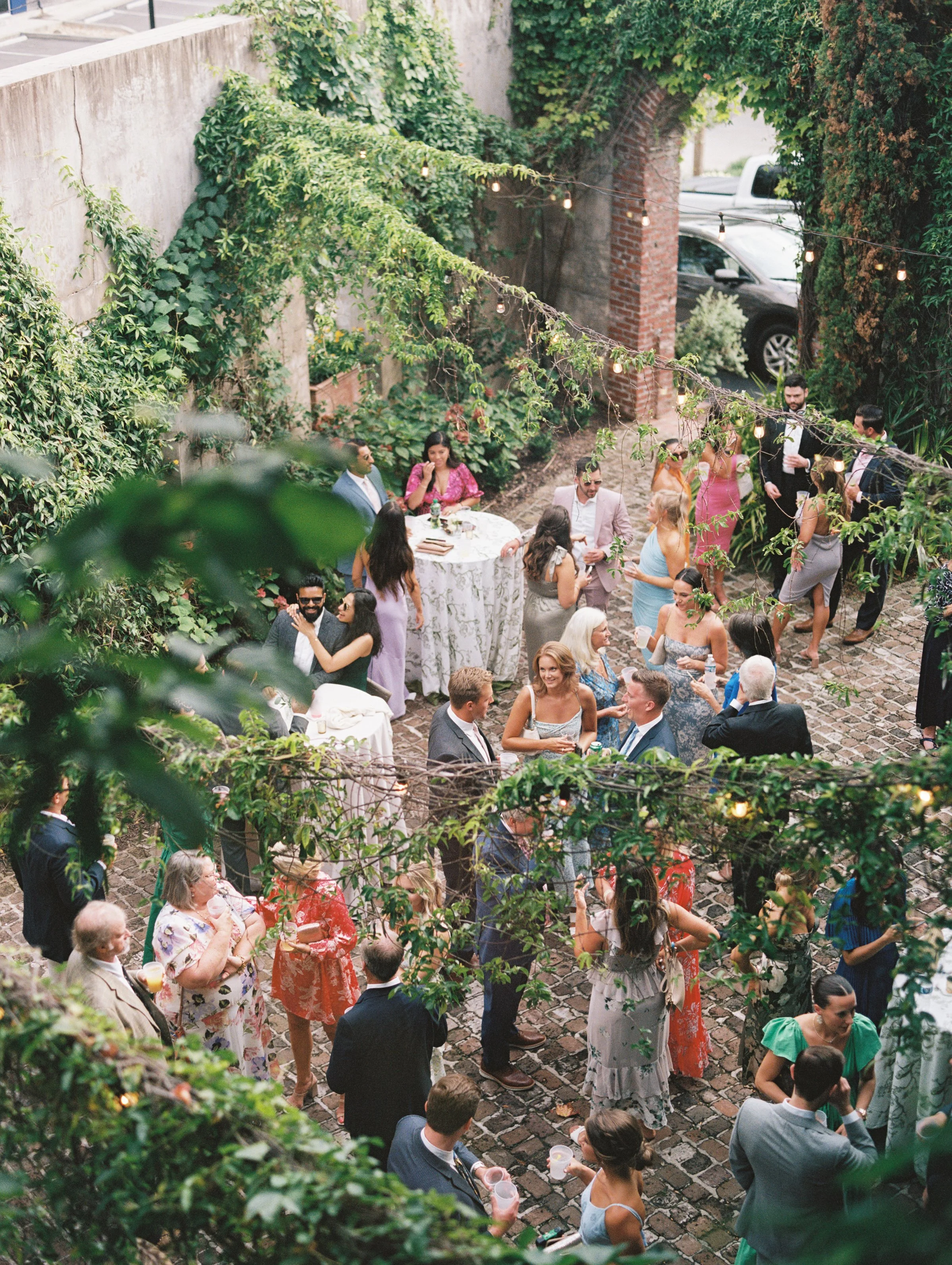 A lively outdoor gathering with guests socializing in a lush garden setting, with string lights overhead and a cobblestone ground.