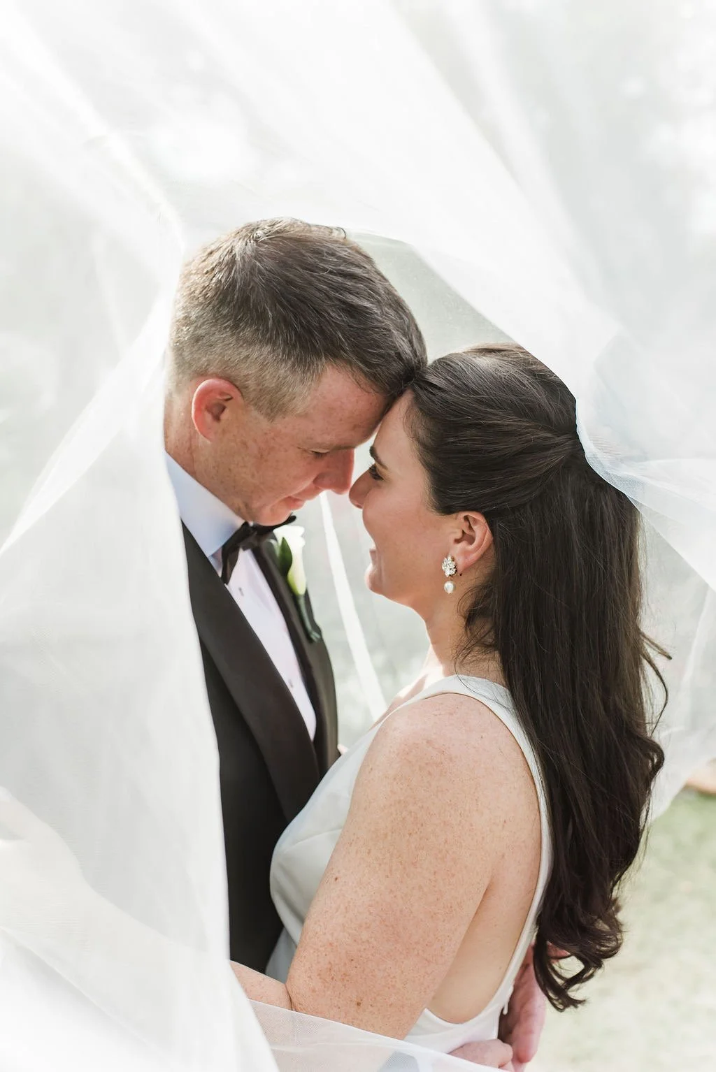 A bride and groom face close with foreheads touching, under a sheer white veil during their wedding.