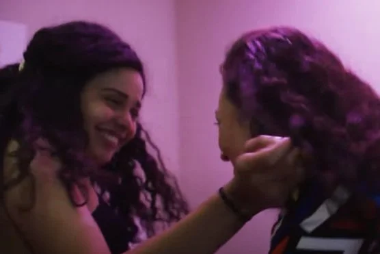 Two gay women smiling and looking at each other in a warmly lit room. One woman is touching the other woman's face while giggling.