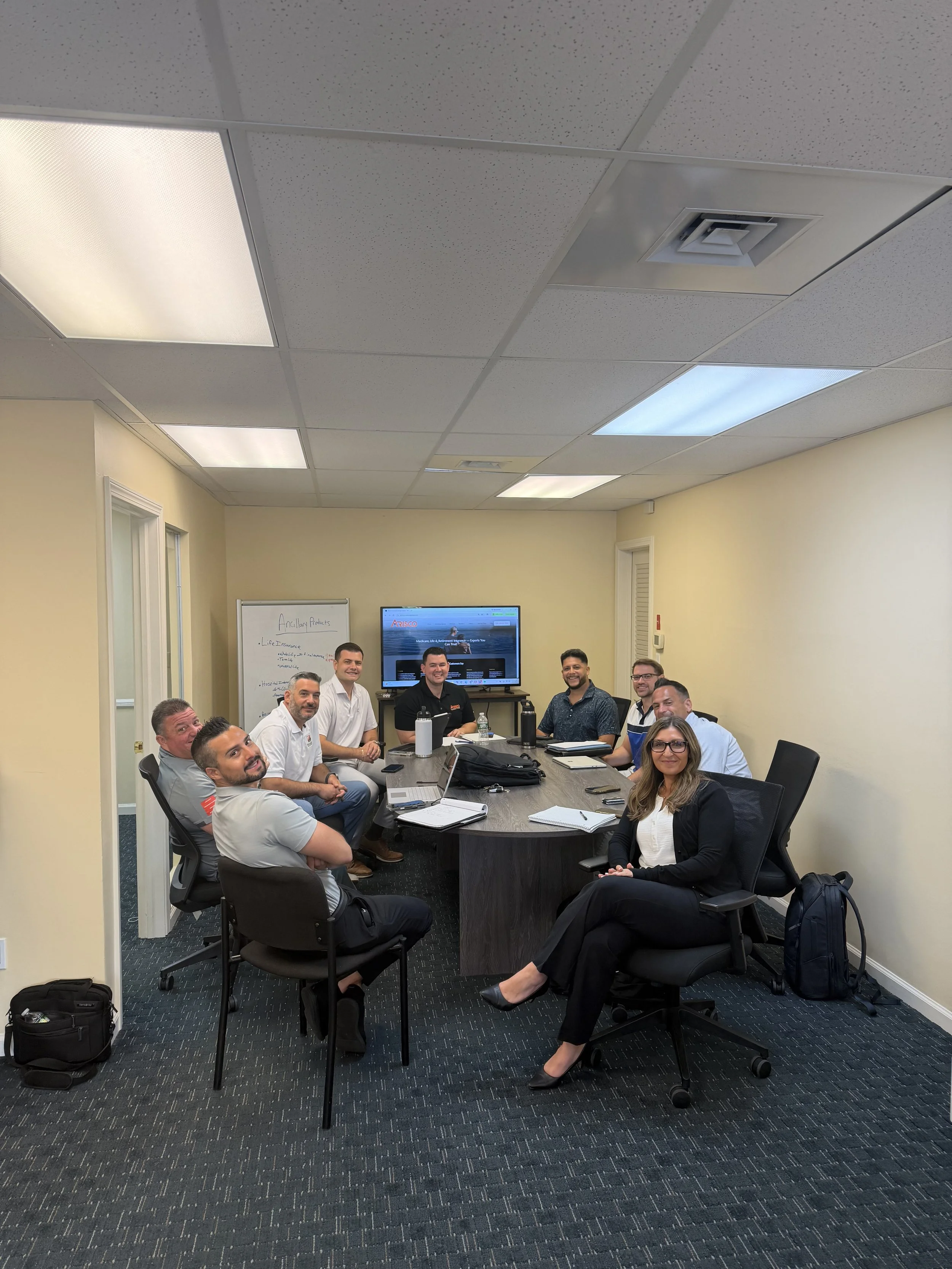 A group of eleven business professionals gathered around a conference table in a meeting room, smiling and looking at the camera. The room has a television monitor, whiteboard, ceiling lights, and cream-colored walls.