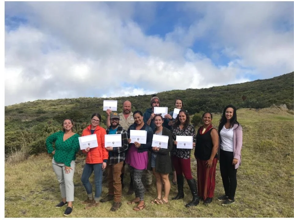 Group of people standing smiling, holding their newly earned training certificates, with green scenery in background with blue cloudy sky.