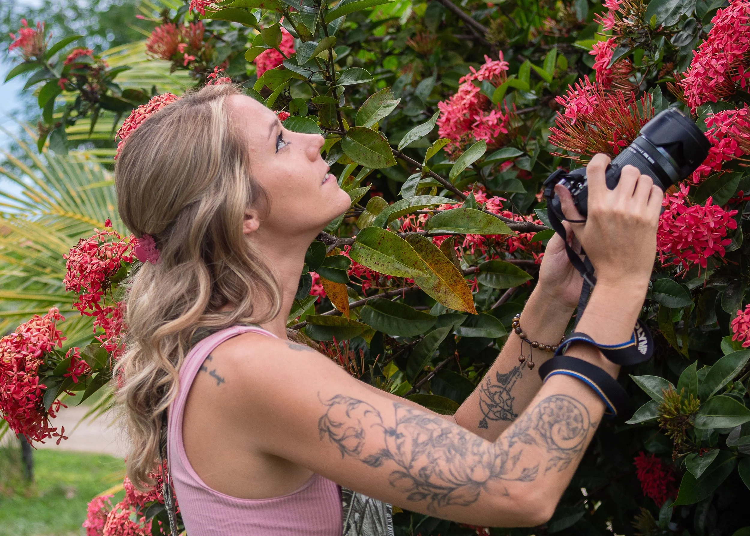 A woman with tattoos on her left arm, wearing a pink sleeveless top, is holding a camera and taking a photo of pink flowers on a bush. She has long wavy blonde hair and is outdoors surrounded by lush greenery.