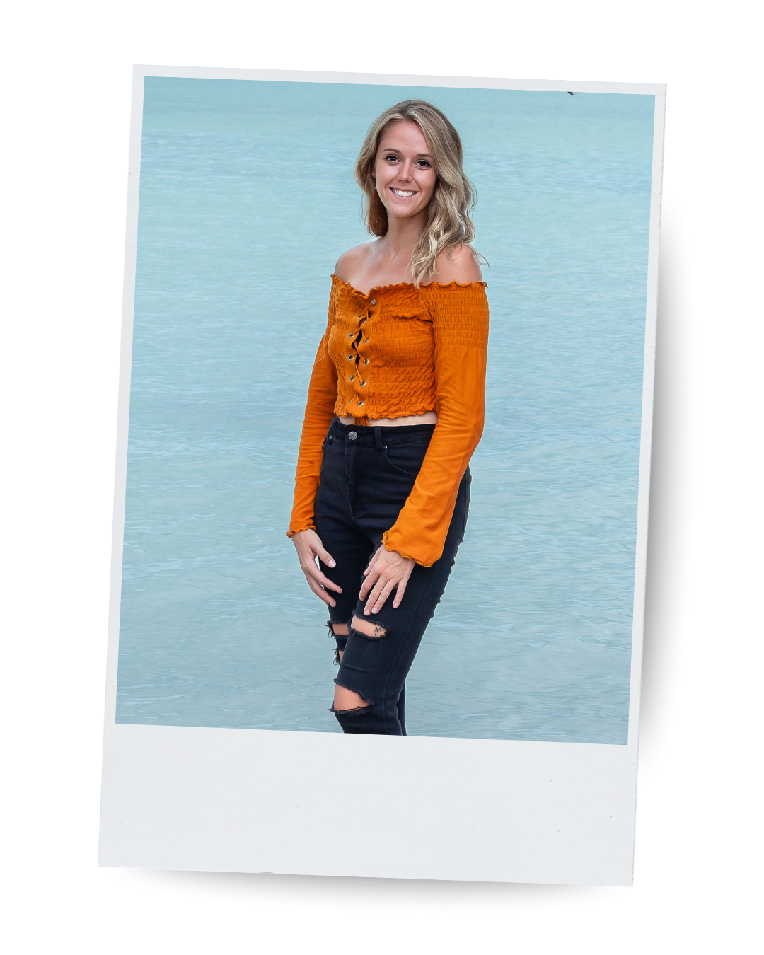 A woman with blonde hair smiling, wearing an orange off-the-shoulder top and ripped black jeans, standing in front of a body of water.