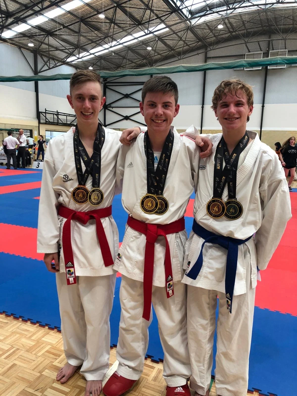 Three young male martial artists in gi uniforms with medals around their necks, standing together on a martial arts competition floor in a gymnasium, smiling.