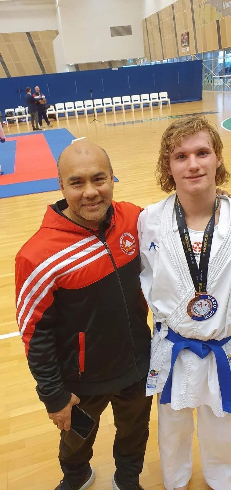A man and a young athlete posing together in a gymnasium, with a martial arts tournament visible in the background. The man is wearing a black and red jacket, and the young athlete is dressed in a white judo gi with a blue belt and medals around his 