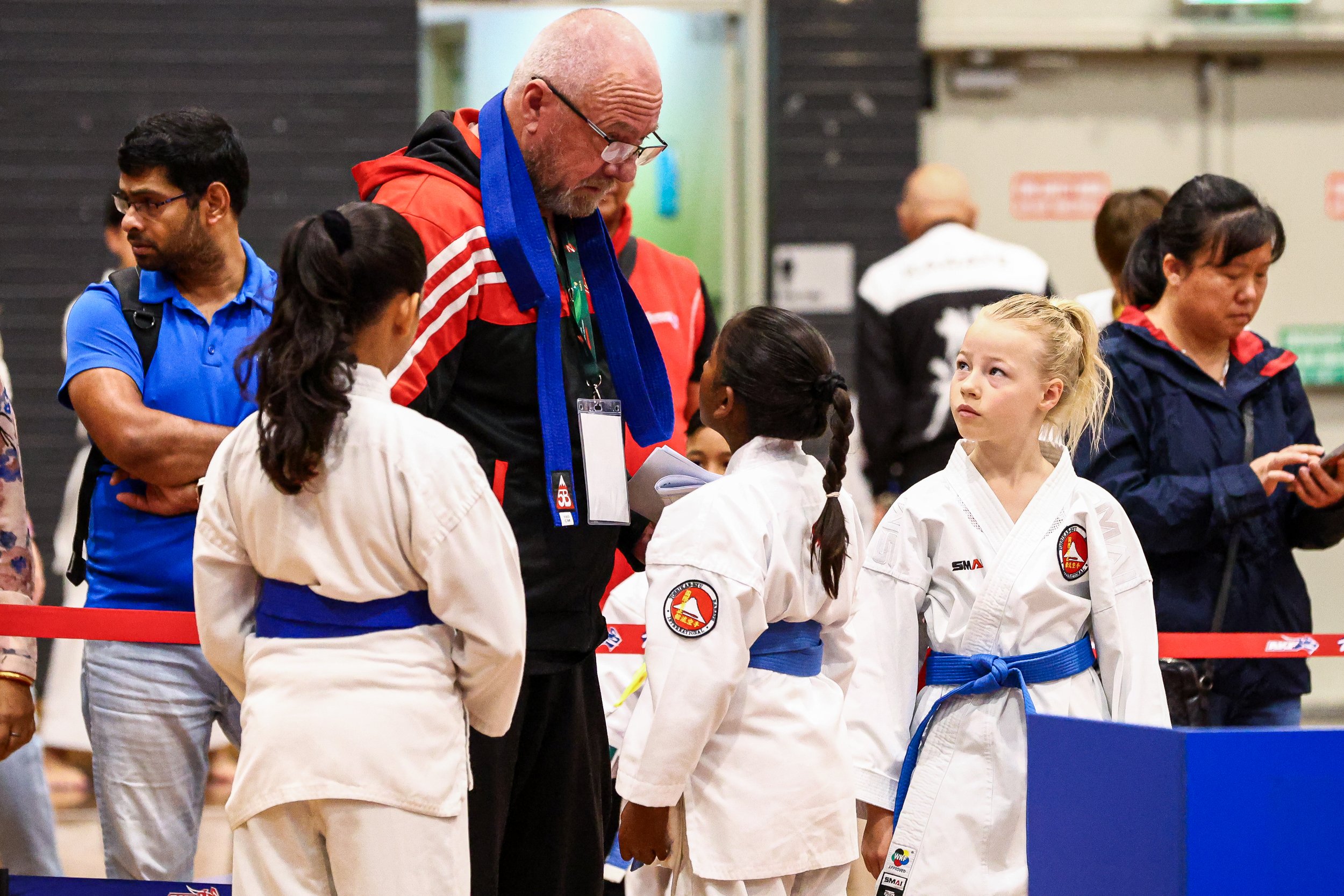 A group of children dressed in white judo uniforms with blue belts, standing in line for a judo event, talking to their coach who is wearing a red and black jacket with a blue scarf, while other adults observe in the background.