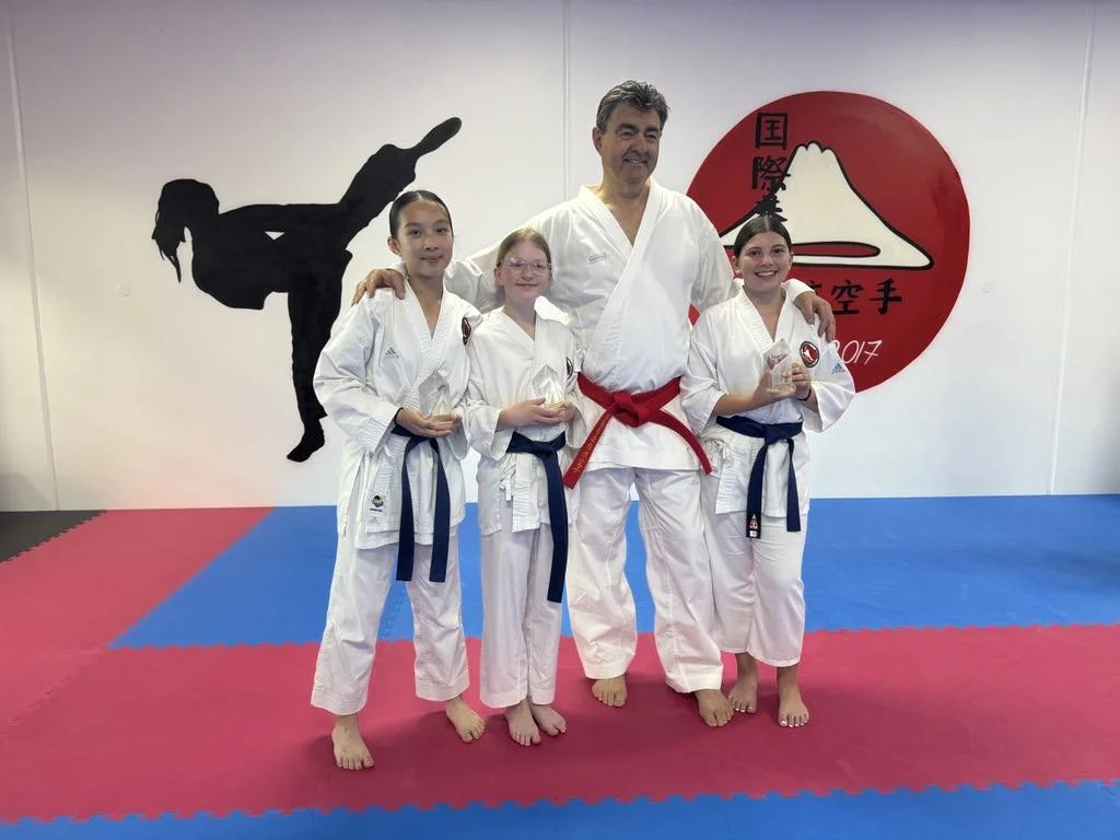 Group of four people in karate uniforms standing on a mat in a dojo, with an adult man in the center and three young girls holding small trophies.
