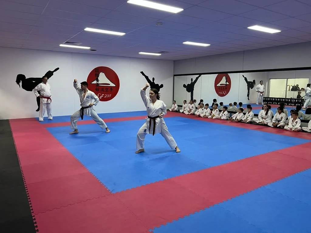 Martial arts class in a dojo with students in white uniforms, some practicing moves, others seated against the wall, and mirrors reflecting the scene.