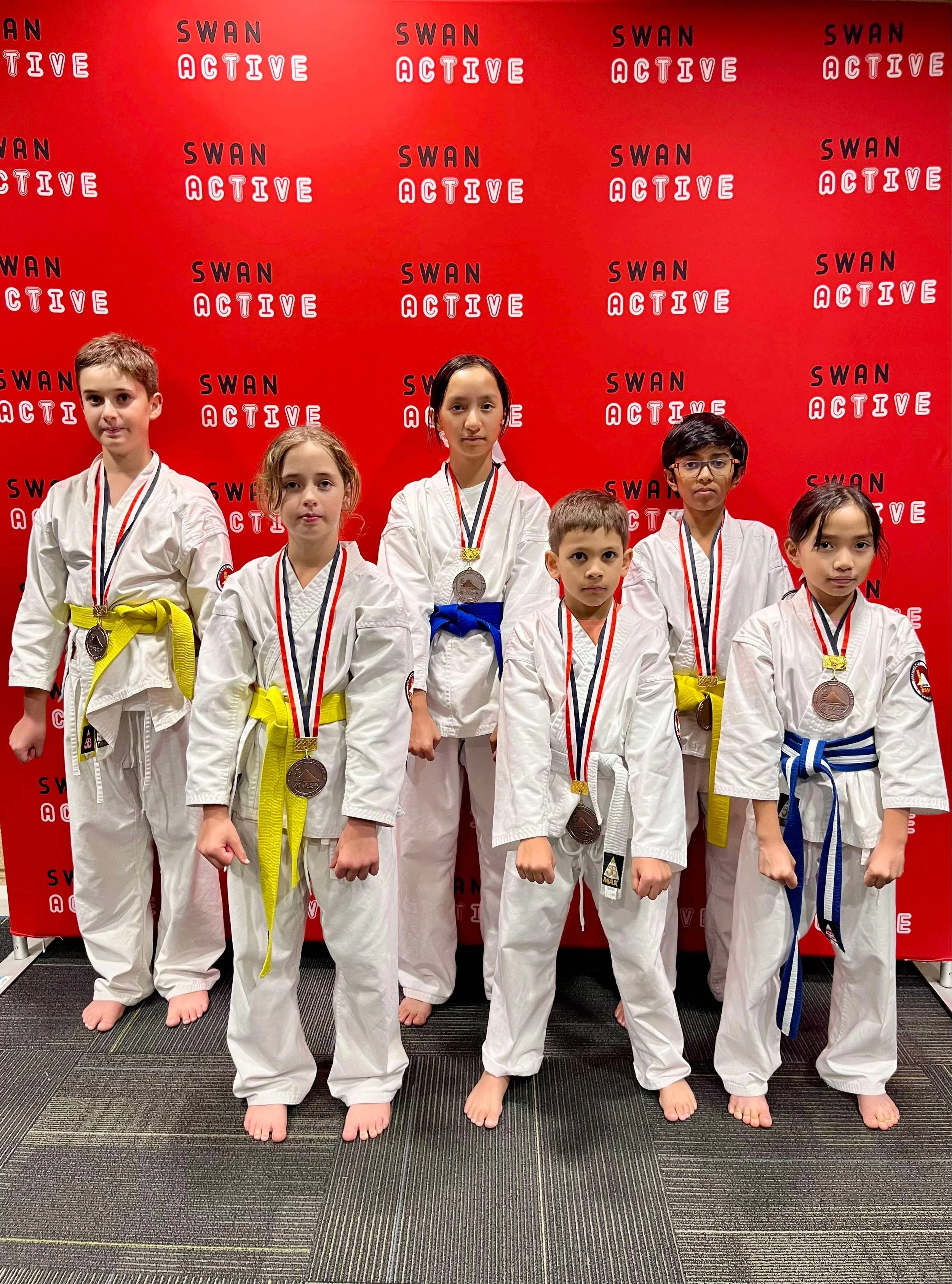 Group of six young martial artists wearing white judogi with various colored belts and medals, standing in front of a red backdrop with 'Swan Active' repeated logo, posing after a competition.