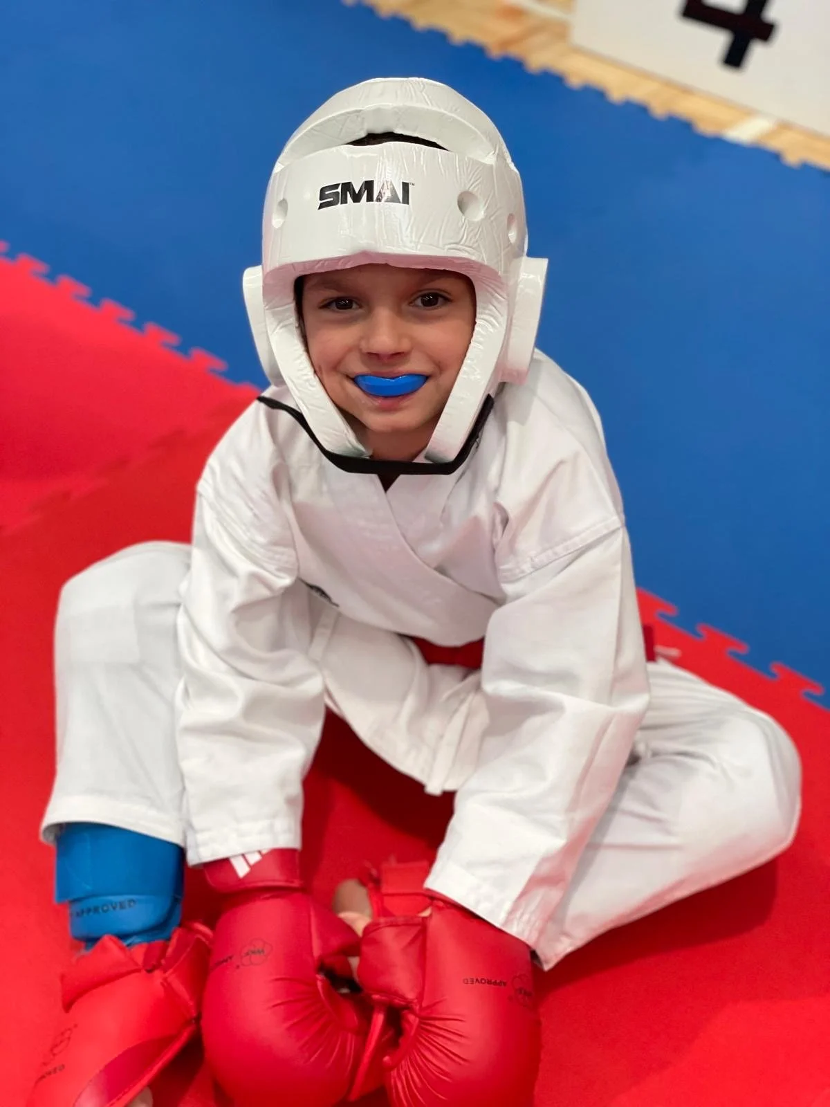 A young boy wearing a white martial arts uniform, a white protective helmet, and red boxing gloves, kneeling on a red and blue padded floor in a martial arts dojo.