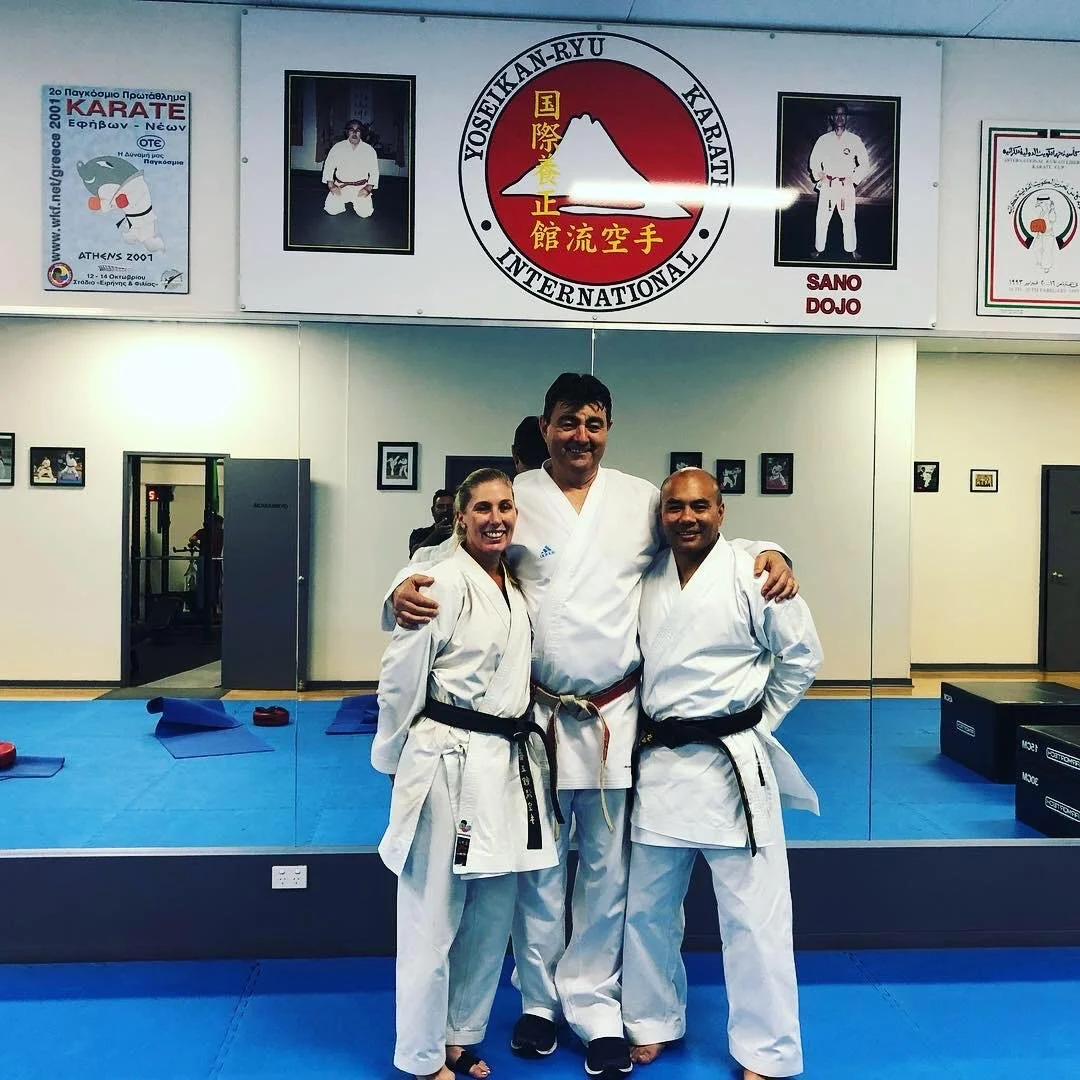 Three martial artists in white gi uniforms with black belts, smiling and posing together in a dojo, standing in front of a mirror and karate-themed posters and banners.