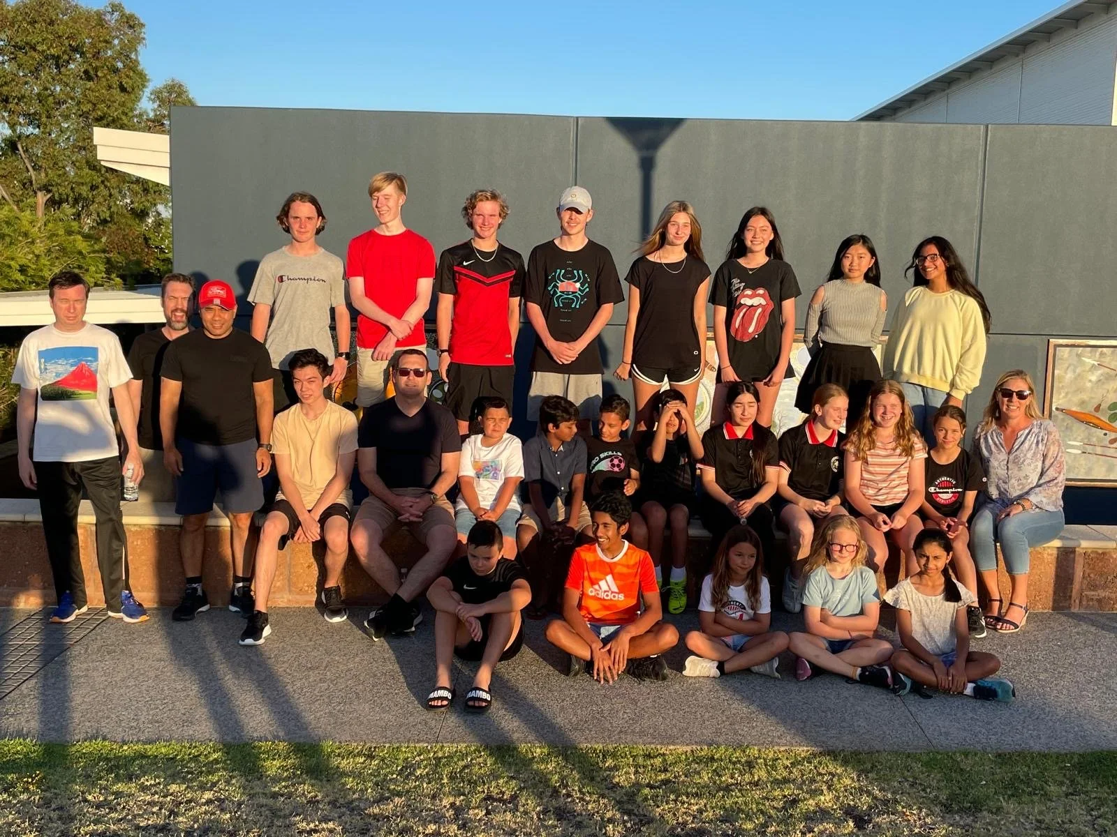 A large group of children and adults posing together outdoors in front of a gray wall during sunset.