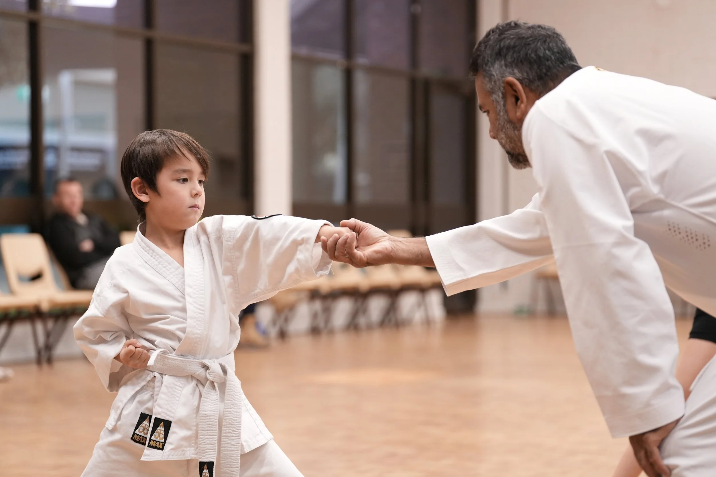 A young boy practicing karate with an instructor in a dojo, with another person sitting on a chair in the background.