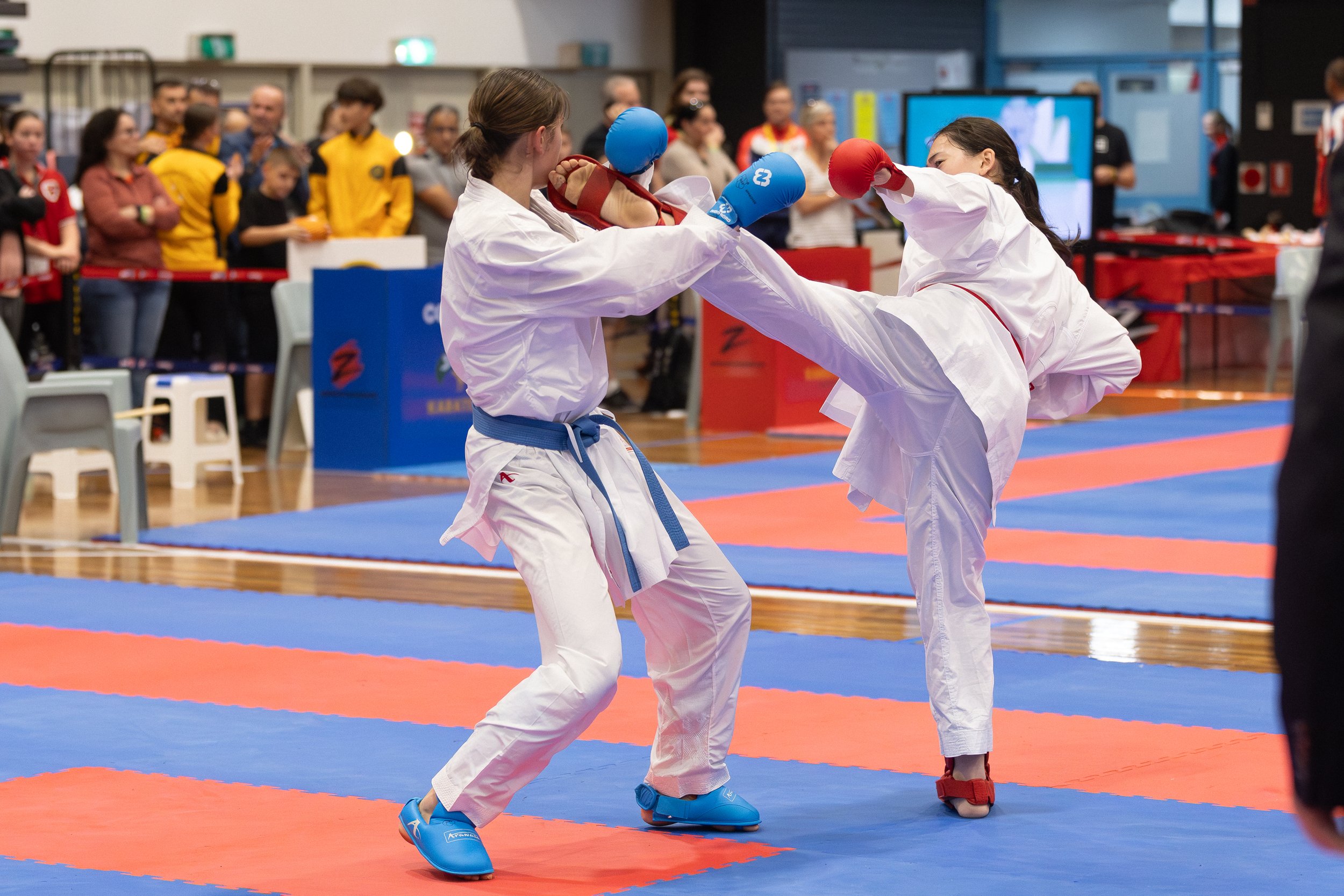Two girls in karate uniforms sparring in a karate match, with one girl delivering a high kick while the other blocks, in a gymnasium filled with spectators.
