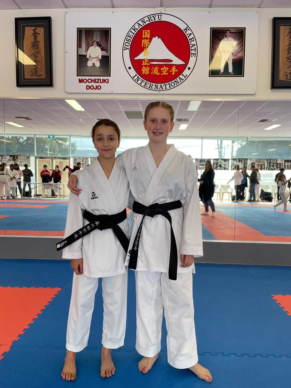 Two young girls in karate uniforms with black belts standing in a martial arts dojo, smiling, with other students and instructors practicing in the background, and banners and photographs on the wall.