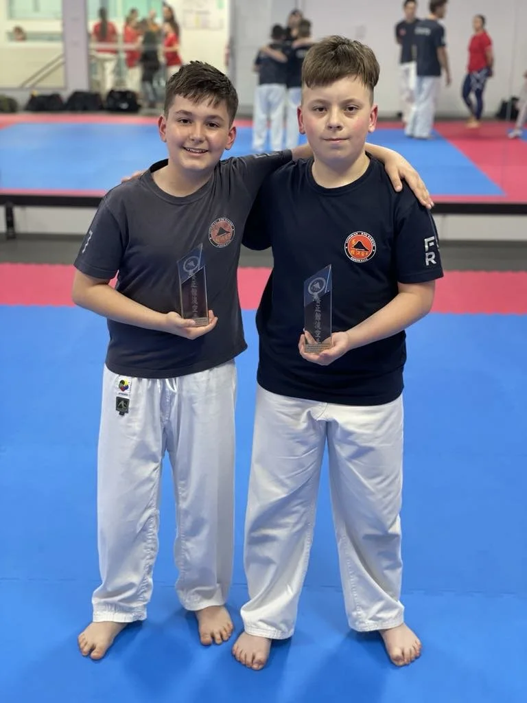 Two young boys in martial arts uniforms standing on a blue mat, holding trophies, with their arms around each other's shoulders, in a martial arts dojo.
