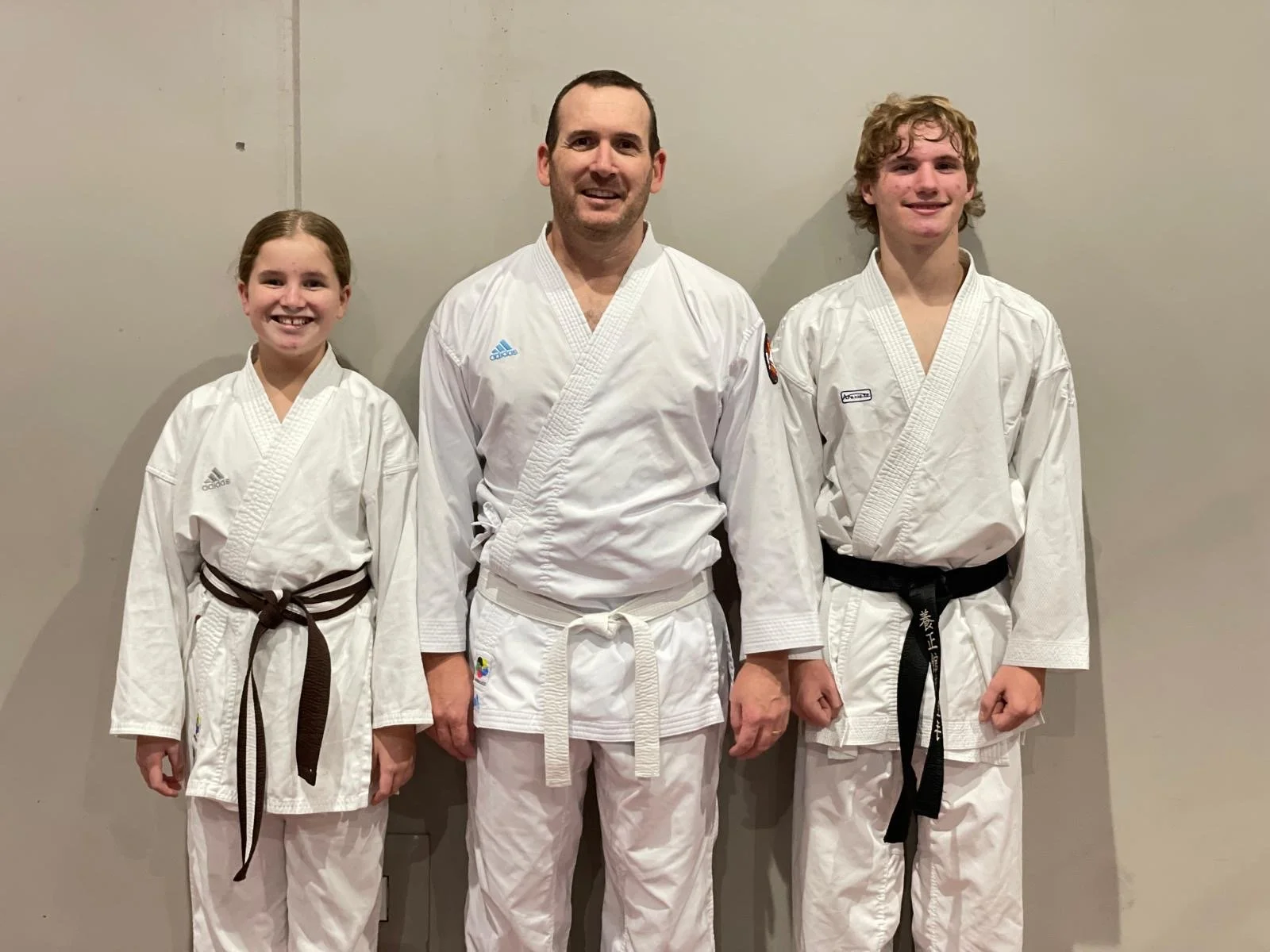 Three martial artists in white gis standing against a plain grey wall, smiling at the camera. The two on the sides are children with brown and blonde hair, and the person in the middle is an adult male with short dark hair.
