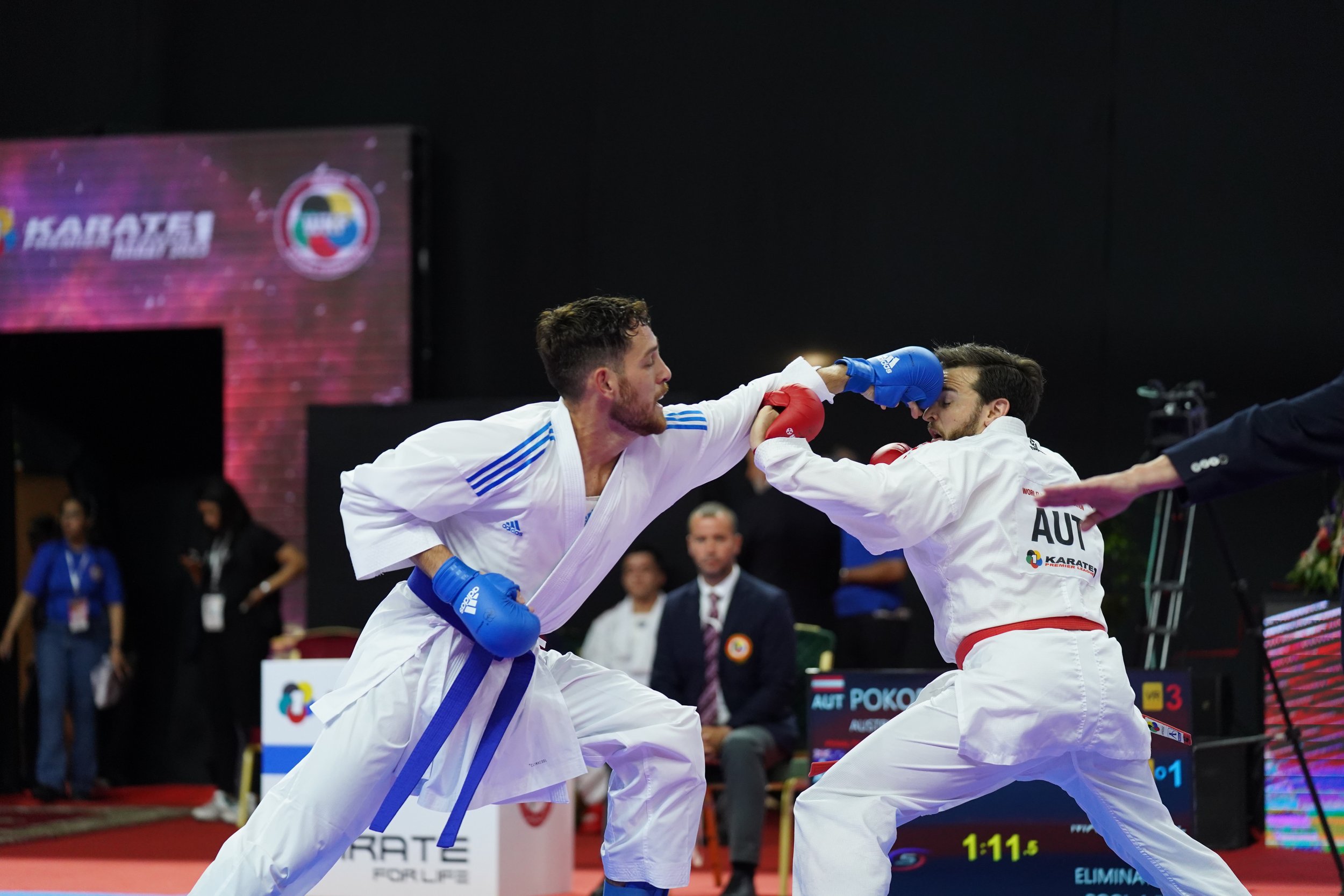 Two male karate competitors in white uniforms sparring, one with blue gloves and the other with red gloves, inside a gym with judges and officials watching.