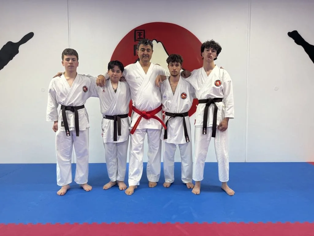 A group of five people dressed in martial arts uniforms, standing on a blue mat in a dojo with a red circle and black markings on the wall behind them. The person in the center has a red belt, and the others have black belts.