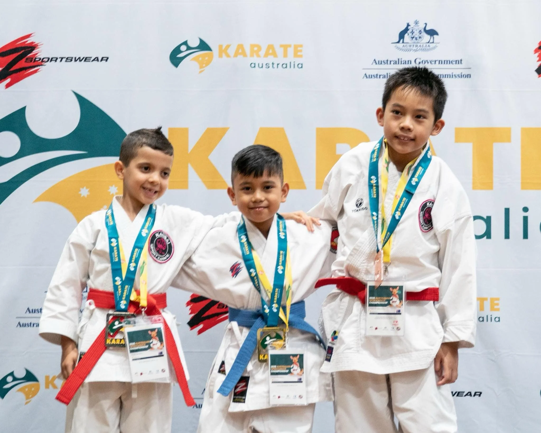 Three young boys in martial arts uniforms standing in front of a backdrop, each wearing medals and smiling after a competition.