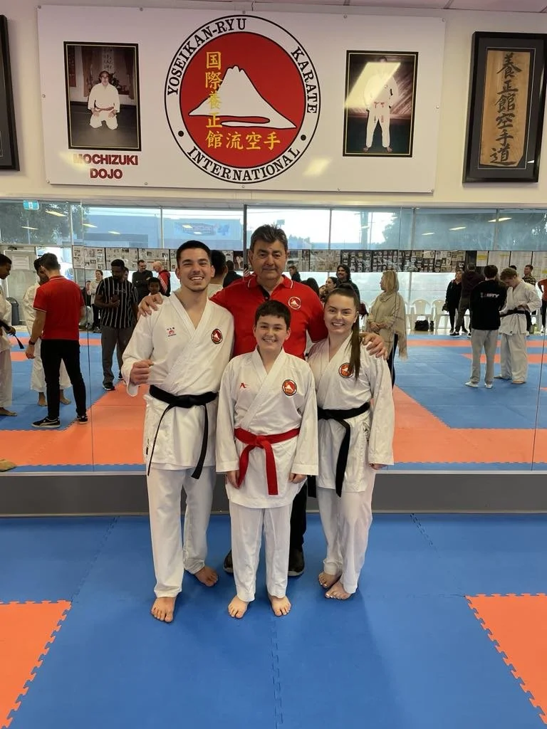 Four people in karate uniforms posing for a photo inside a karate dojo, with a man in a red shirt and a group of people training in the background.