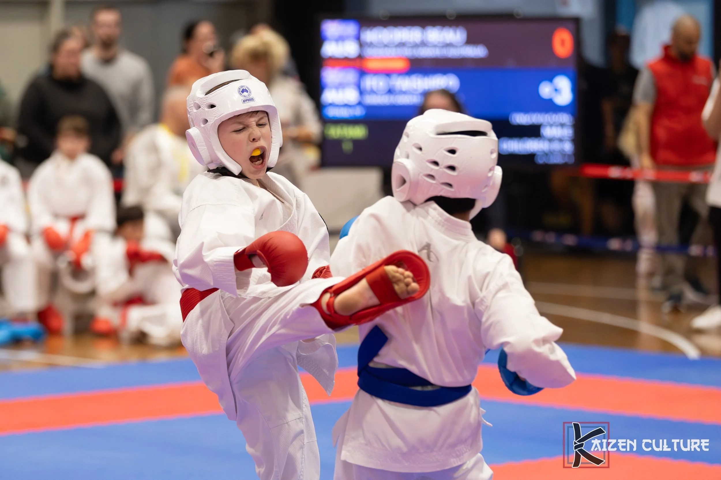 Young martial artists sparring in a karate competition, wearing protective helmets and gloves, with spectators watching in the background.
