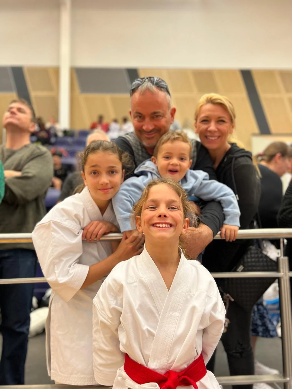 A smiling family with children in judo uniforms posing together in an indoor sports arena.