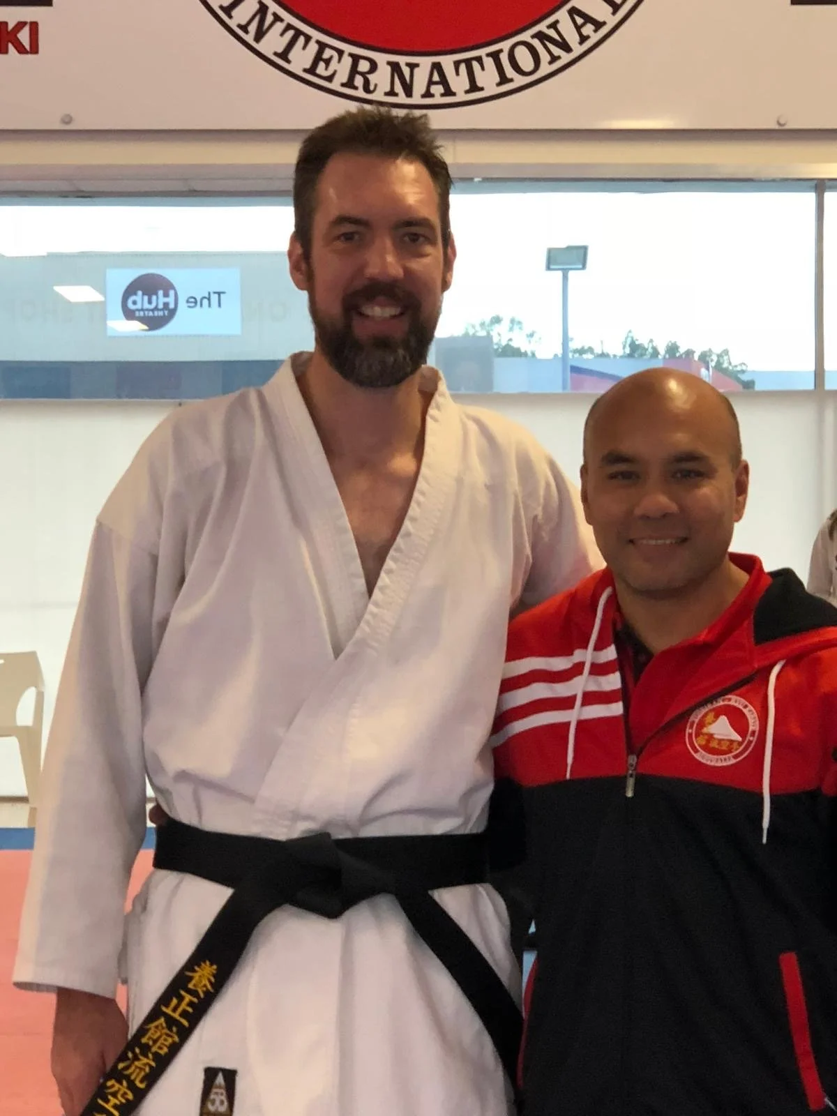 Two men smiling, one in a white martial arts uniform with a black belt, and the other in a red and black sports jacket, standing inside a martial arts gym.