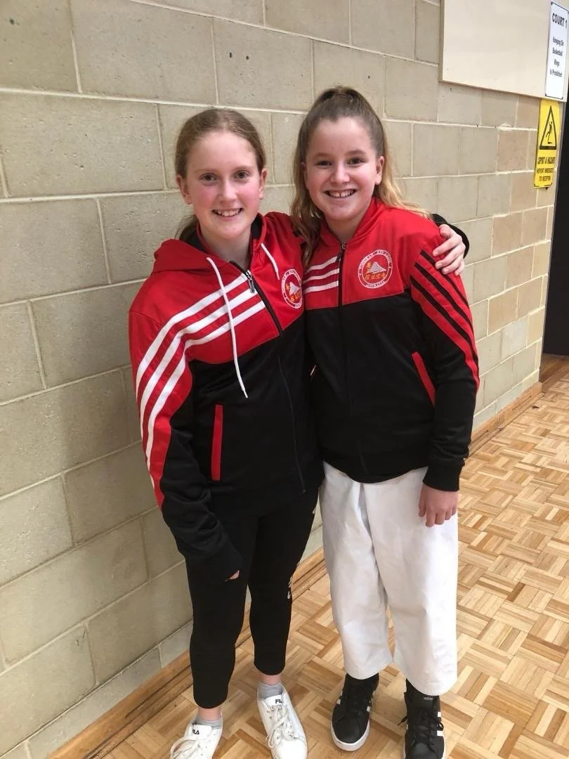 Two young girls with long hair, wearing matching red and black sports jackets, smiling and hugging each other in a gymnasium hallway.