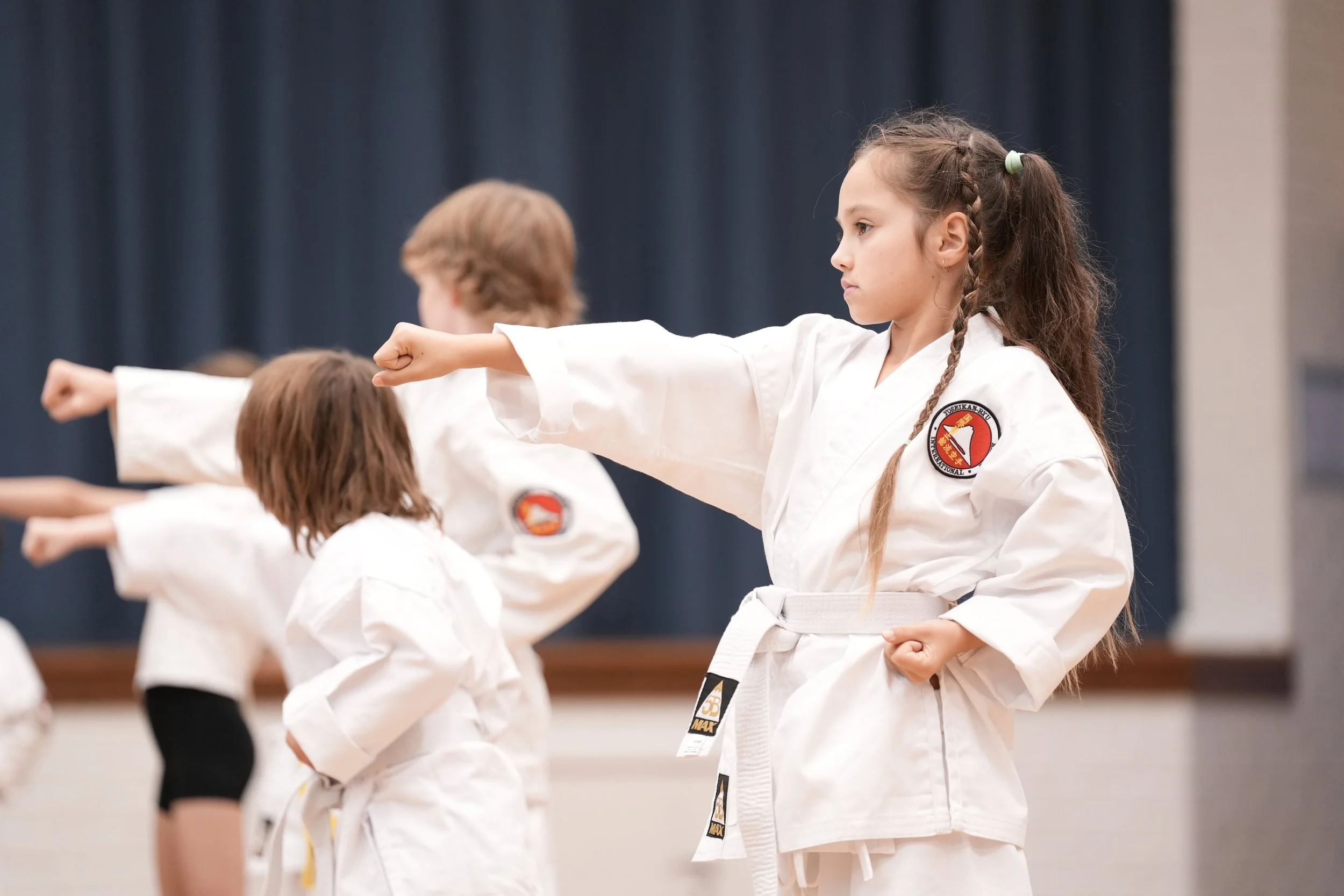 Young girl practicing martial arts in a uniform with a patch, performing a punch during a class with other children.
