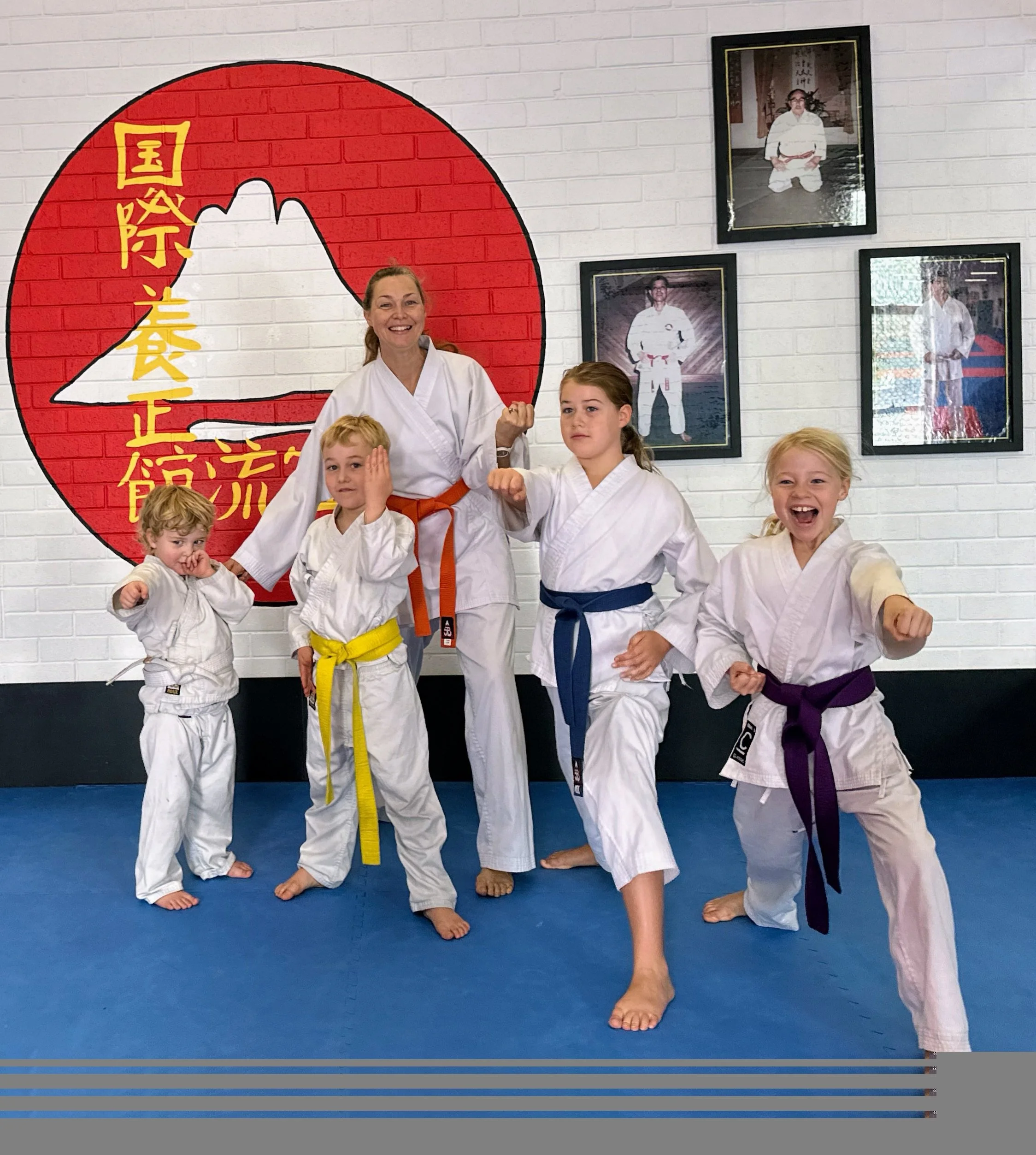 A group of five children and their instructor in a karate dojo, wearing karate uniforms with colored belts, posing in martial arts stances in front of a wall with a Japanese flag and framed photos.