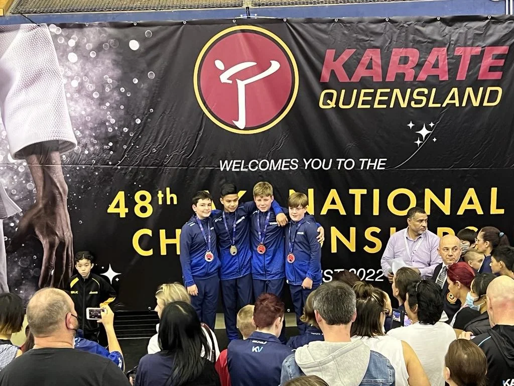 Young athletes standing on a podium with medals at the 48th Karate Queensland Championship in 2022, surrounded by spectators and officials.