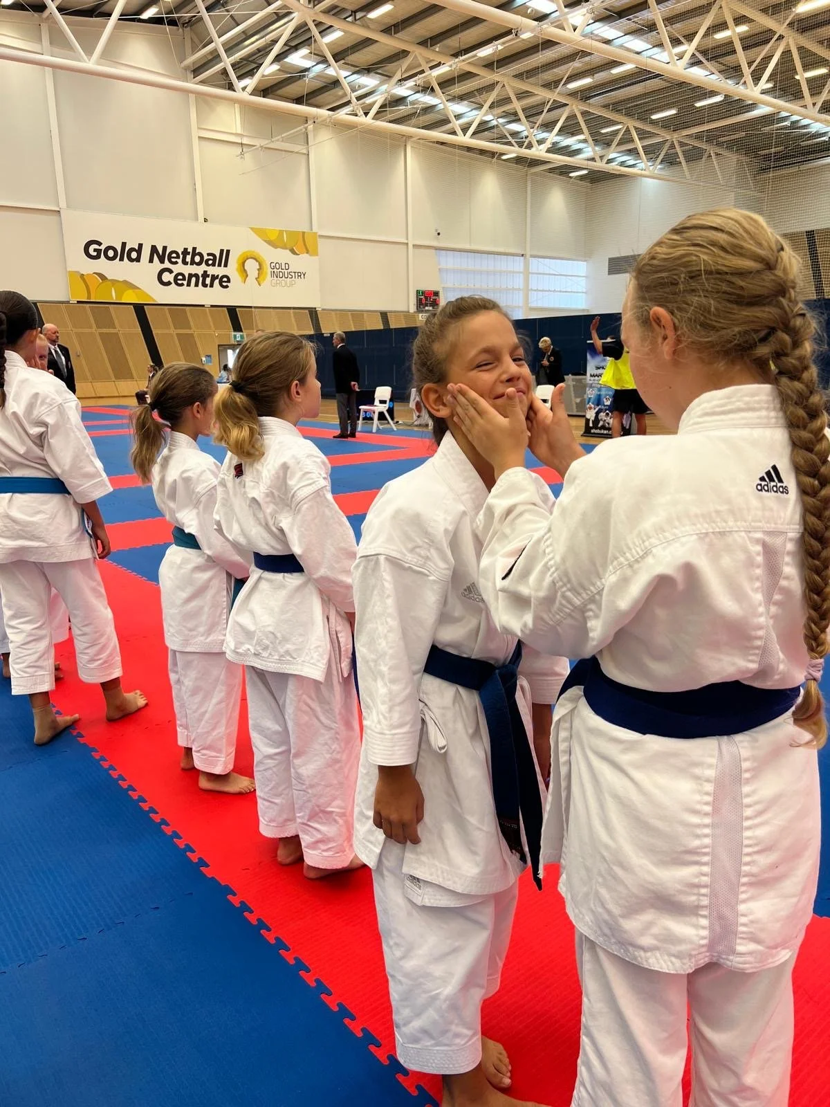 Children in judo uniforms lined up in a dojo, one girl receiving a rub on her cheeks from another girl, with a sign that reads "Gold Netball Centre" in the background.